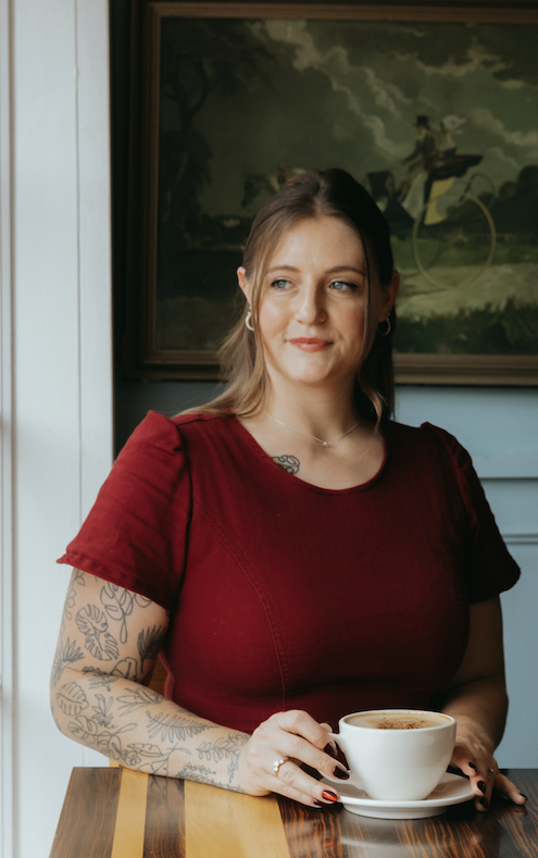 A woman with tattoos on her arm sitting at a table, holding a cup of coffee, wearing a red shirt, with a painting in the background.