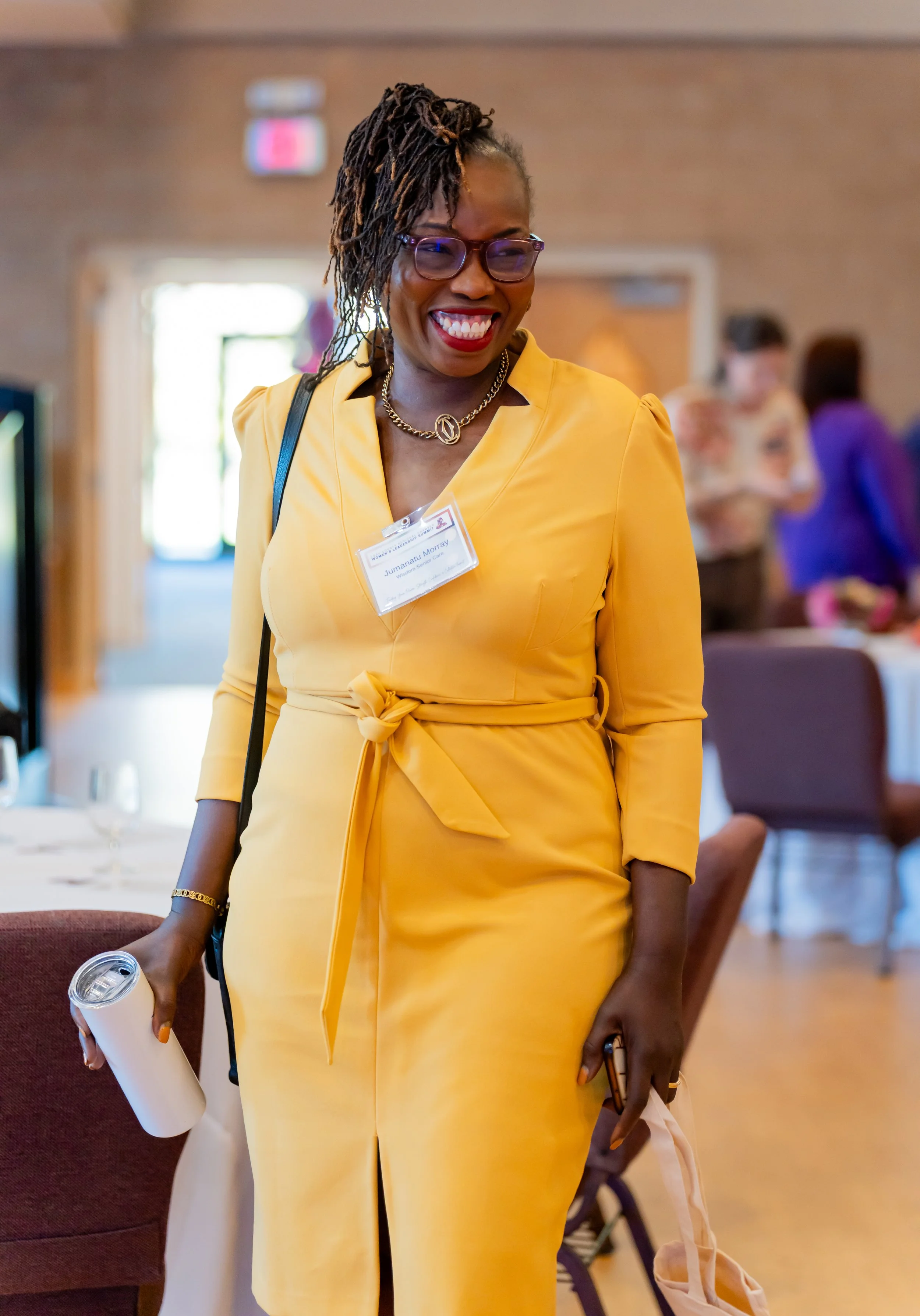 A woman in a yellow dress with a name tag, smiling at a social event in a indoor venue.