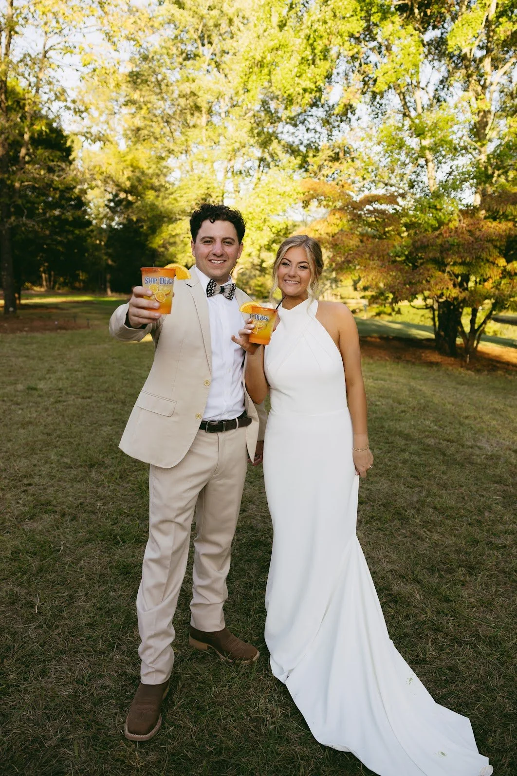 A wedding couple in a park holding drinks with orange slices, trees in the background.