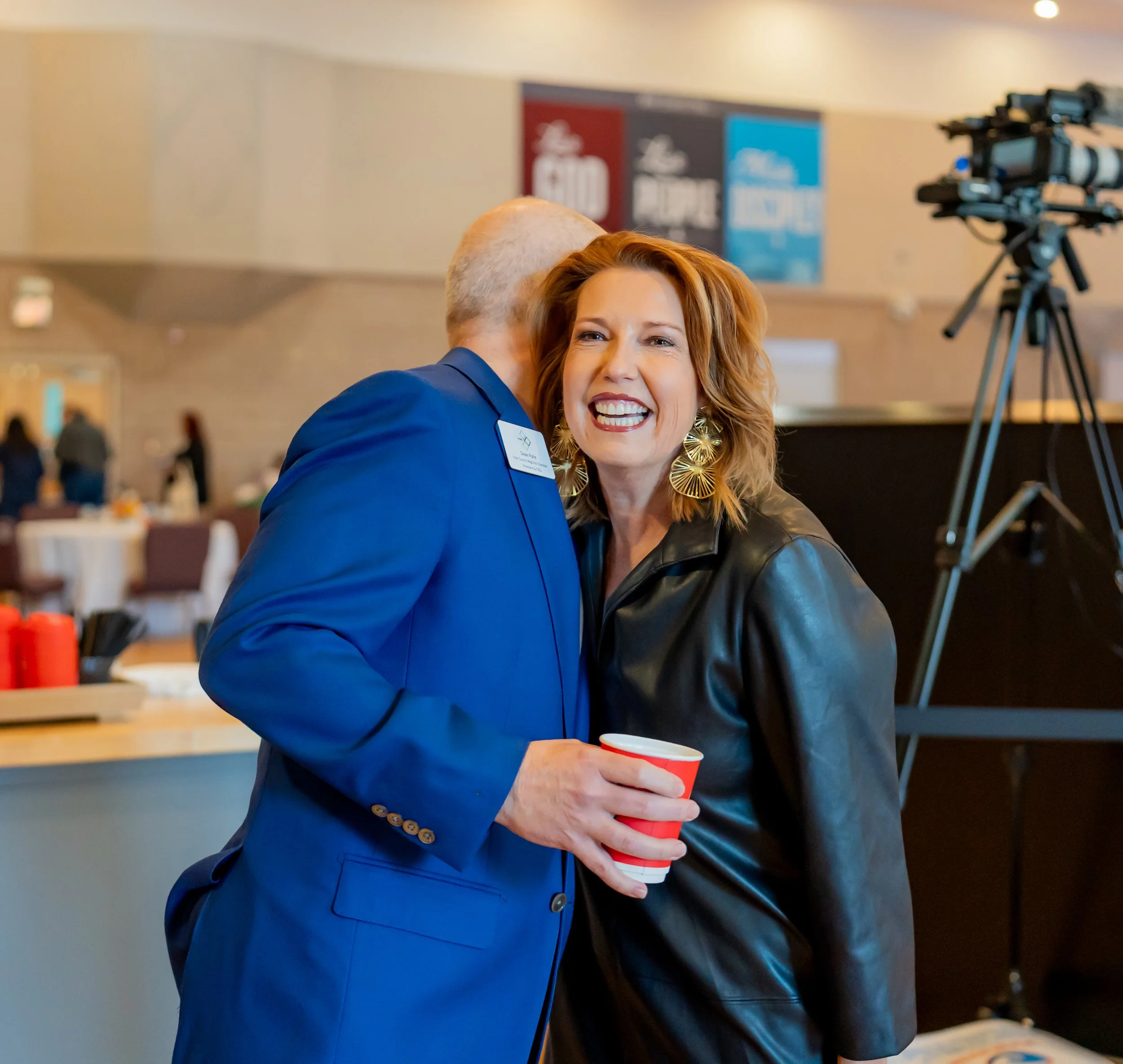 Two people sharing a friendly hug at an indoor event, one holding a red cup, with a camera and banners in the background.