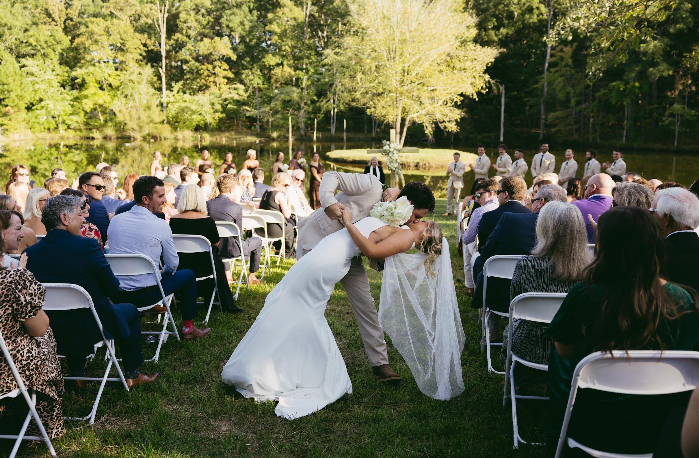 A couple sharing a kiss during their outdoor wedding ceremony by a lake surrounded by trees, with guests seated on white chairs.
