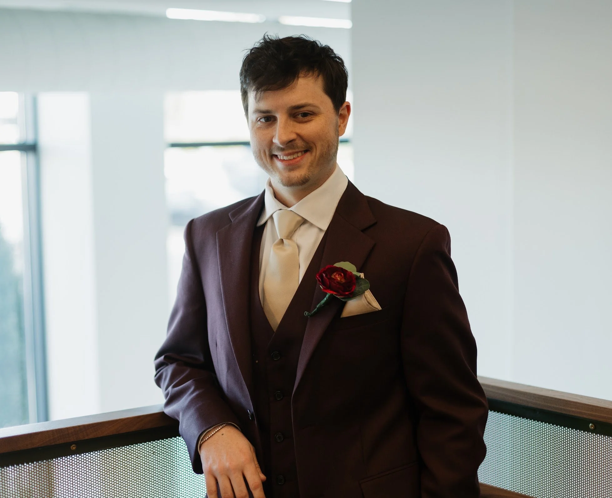 A man wearing a maroon suit with a cream-colored tie and a red boutonniere, smiling indoors near a wooden barrier.