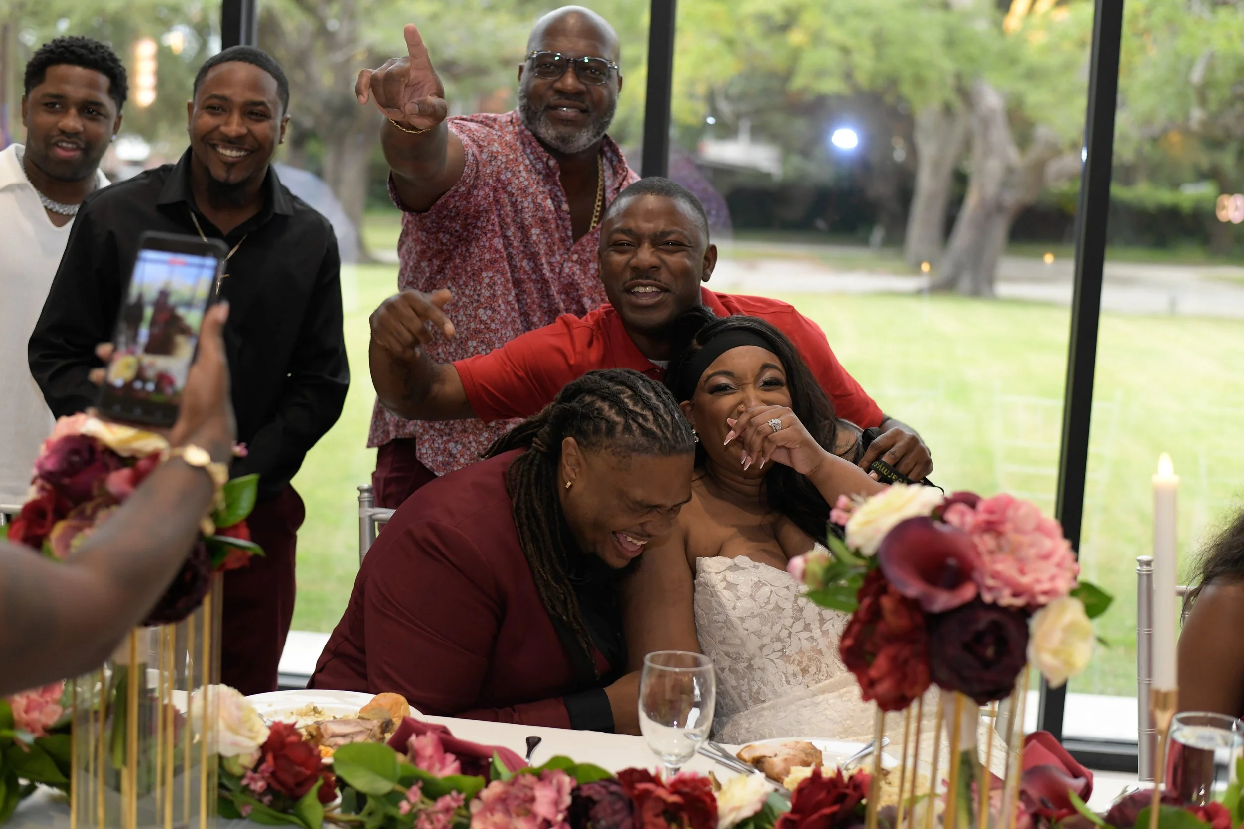 A group of friends celebrating at a wedding, sitting around a table with flowers, with some standing and taking photos, and everyone smiling and laughing.