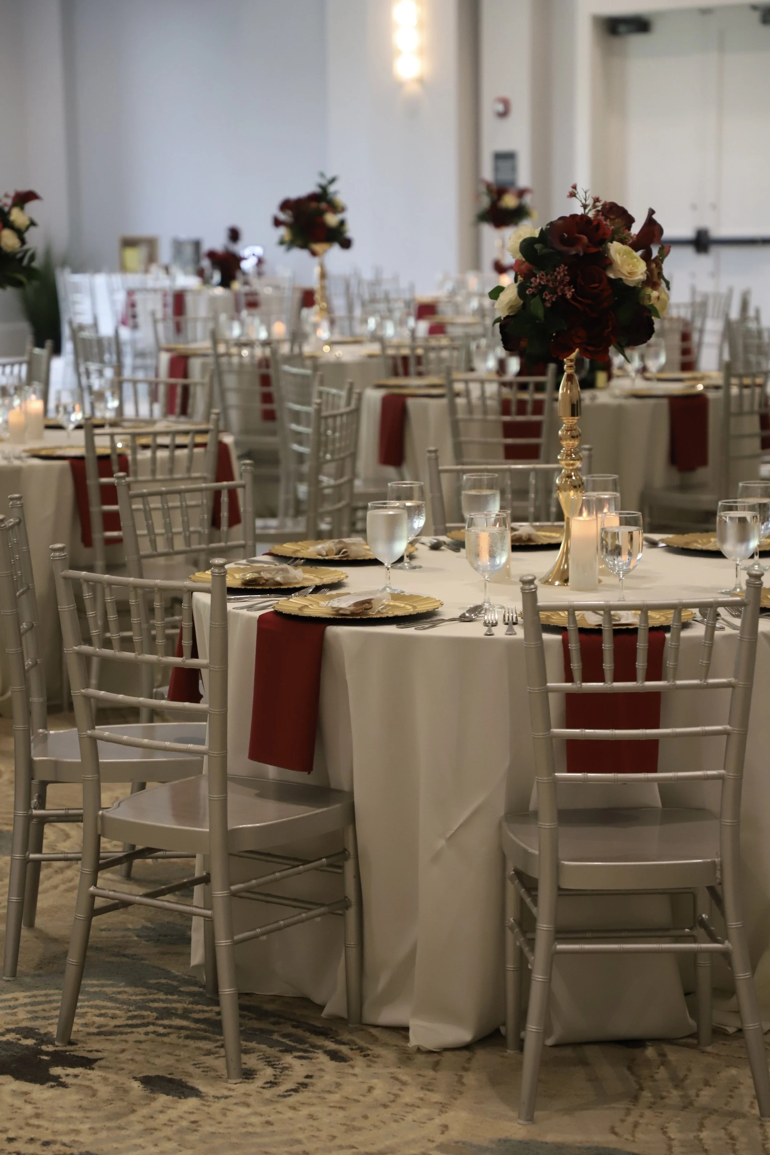 Elegant banquet table with floral centerpiece, gold accents, and red napkins in a well-lit event hall.