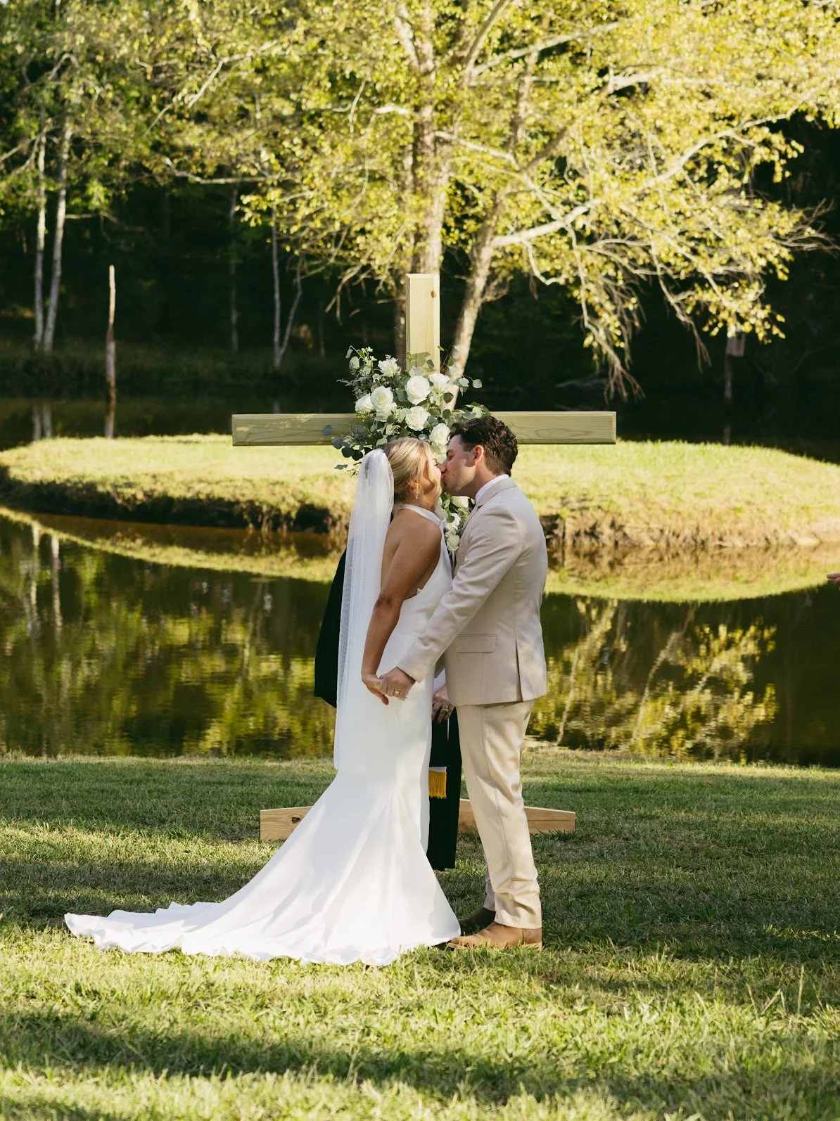 A bride and groom kiss in front of a wooden cross decorated with white flowers and greenery, near a pond with trees in the background on a sunny day.