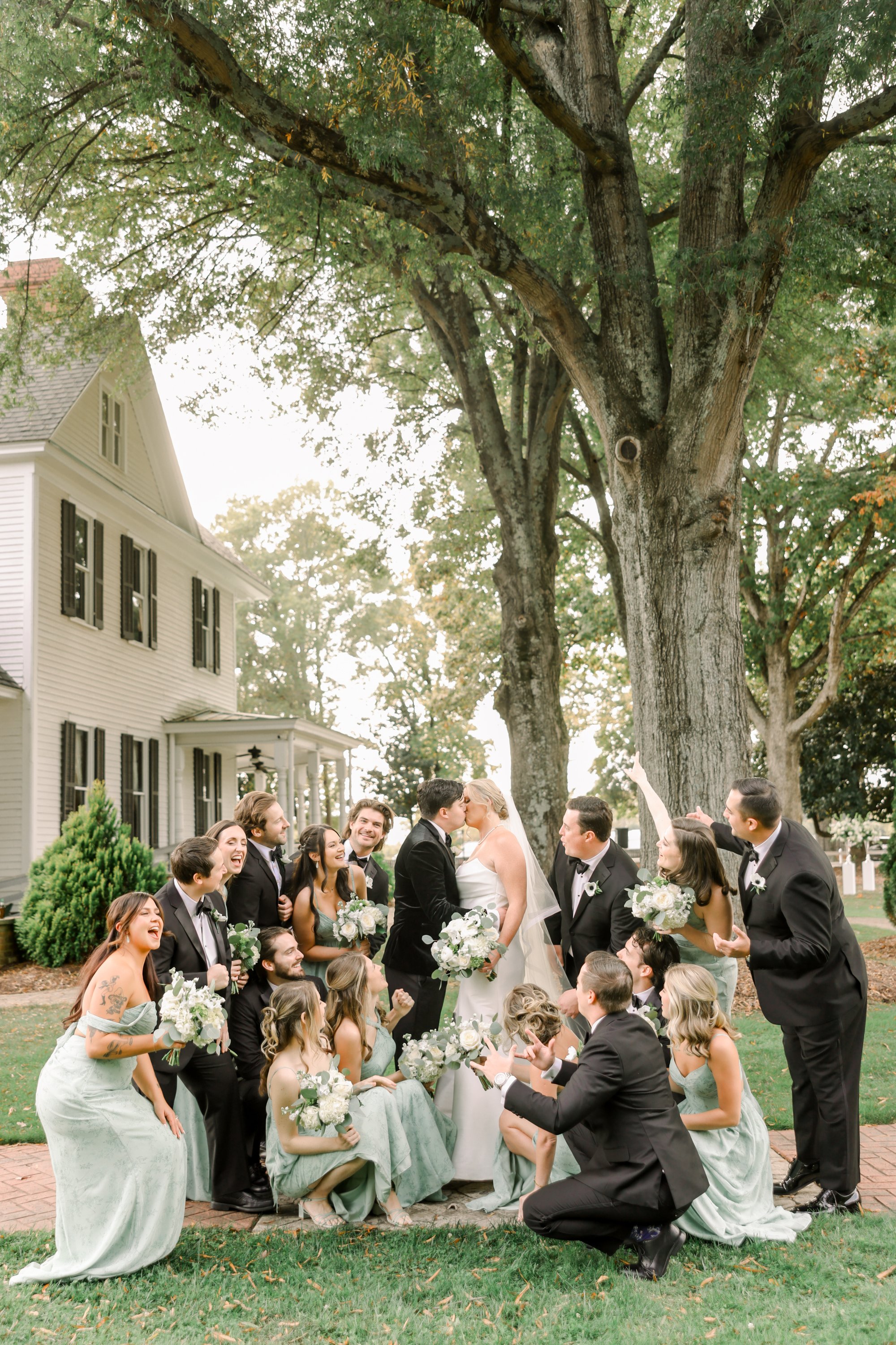 A wedding group photo featuring a bride and groom kissing under a large tree, surrounded by bridesmaids and groomsmen in formal attire, outdoors in front of a white house with black shutters.