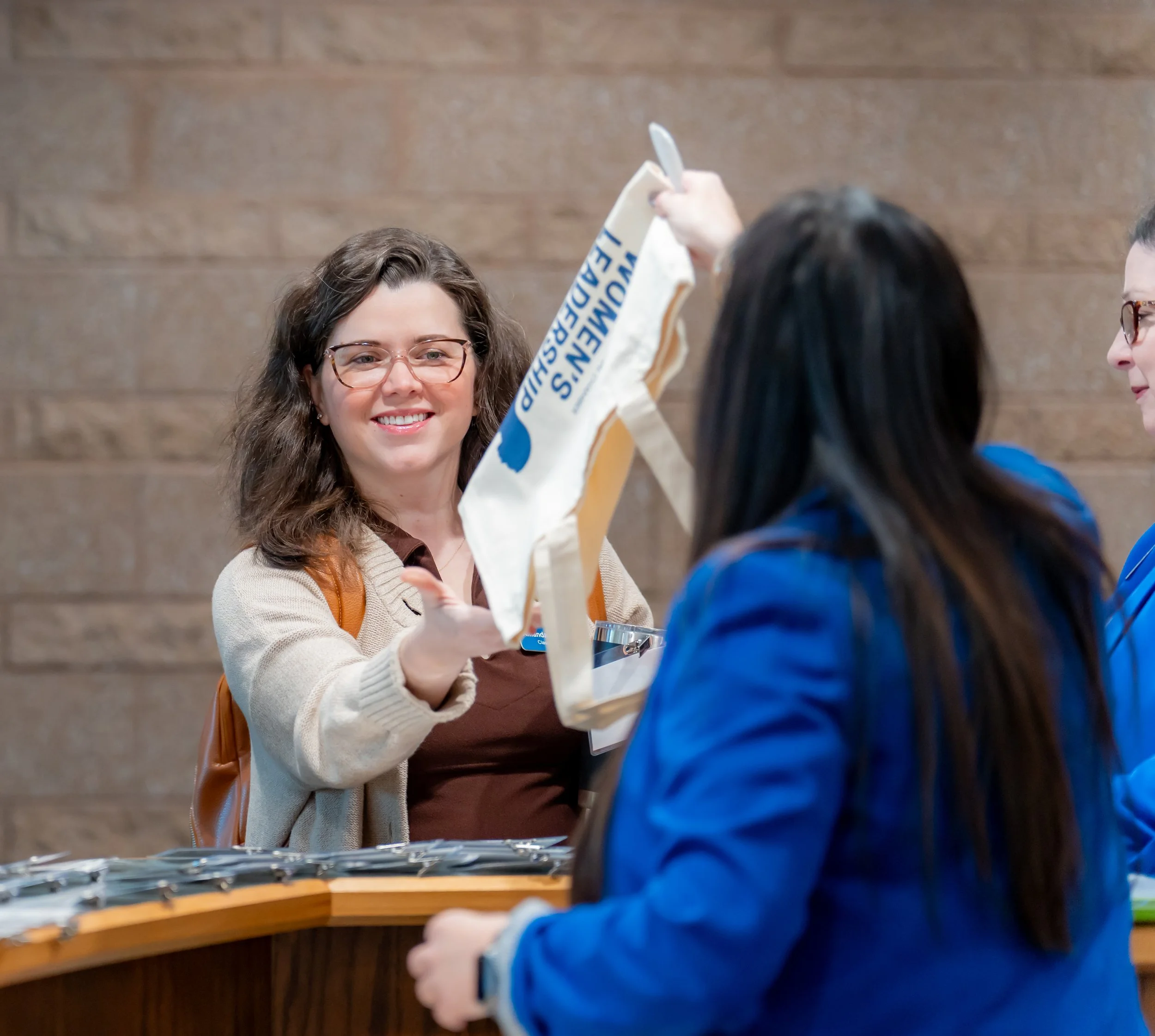 A woman with glasses and curly brown hair receiving a tote bag from another woman wearing a blue jacket at a registration table.