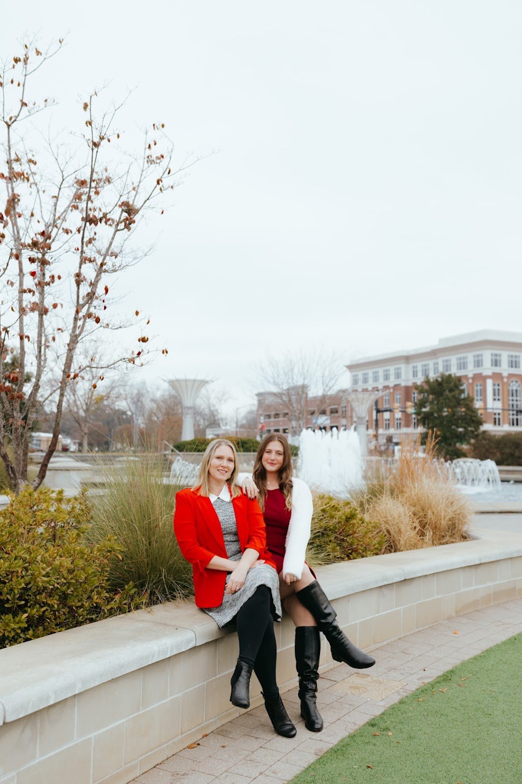 Two women sitting on a stone ledge near a fountain, dressed in fall or winter clothing, with a water tower and buildings in the background.