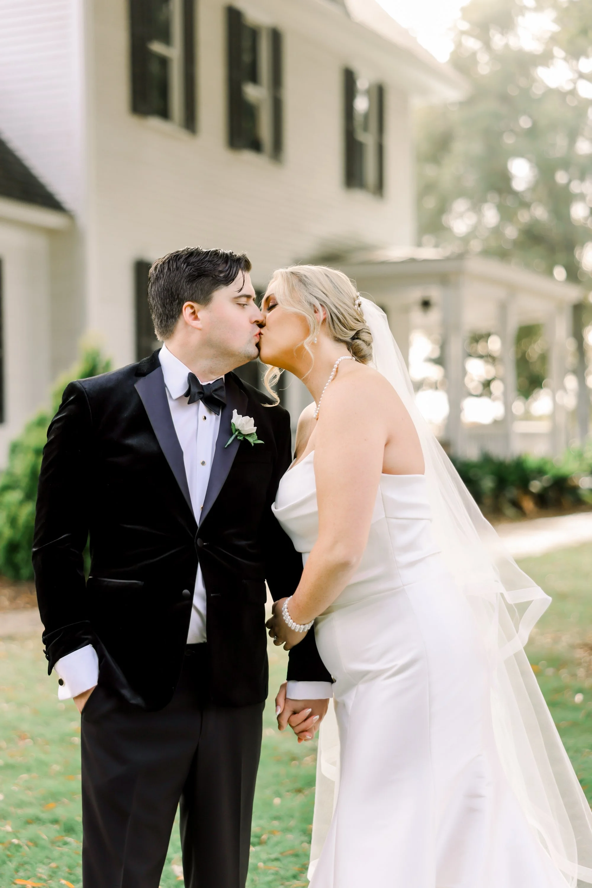 A newlywed couple shares a kiss outdoors in front of a house, dressed in wedding attire with the groom in a black tuxedo and the bride in a white gown with a veil.