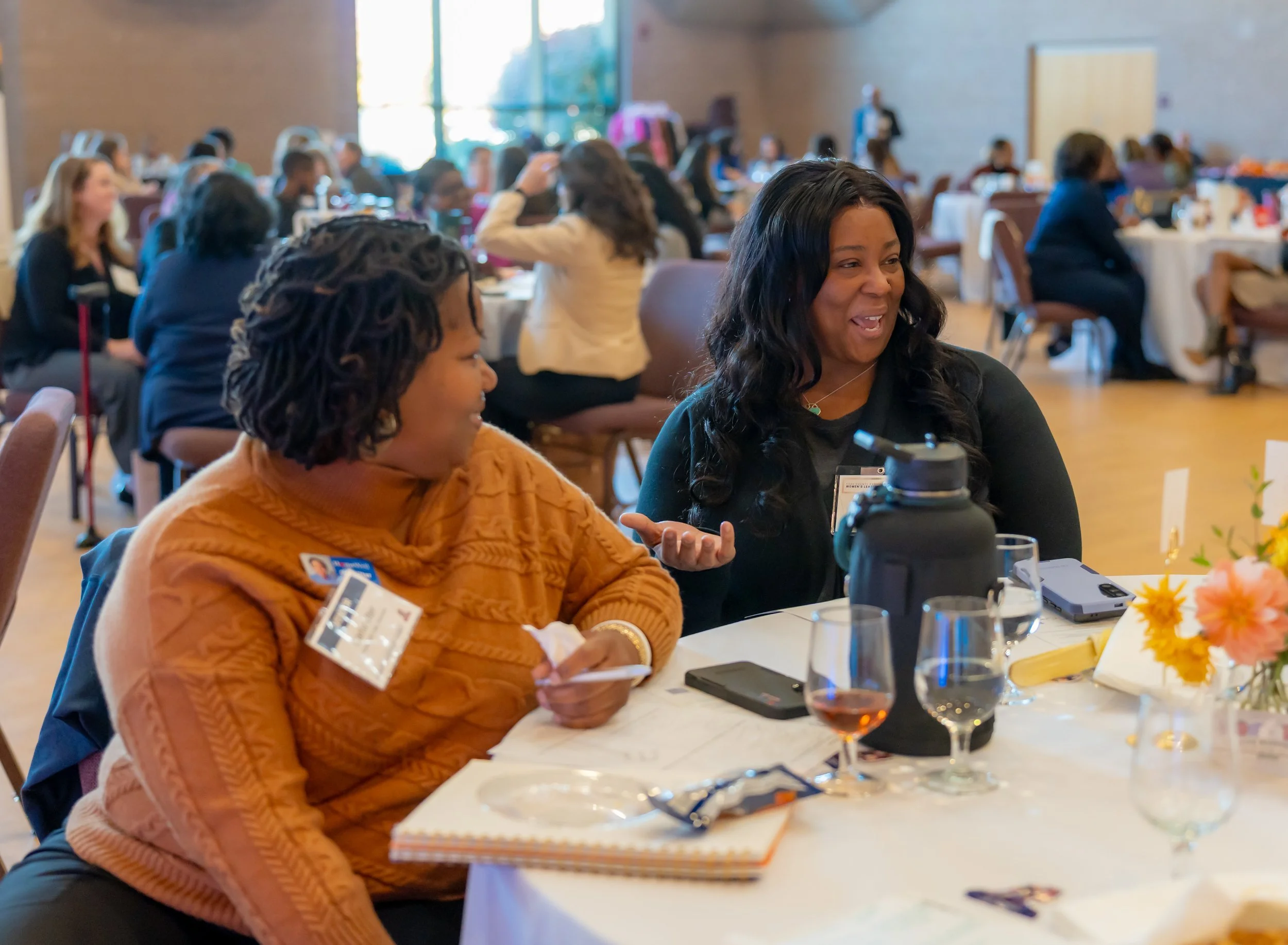 Women sitting at a round table during a conference or event, engaging in conversation. Other attendees are visible in the background in a large room with natural light.