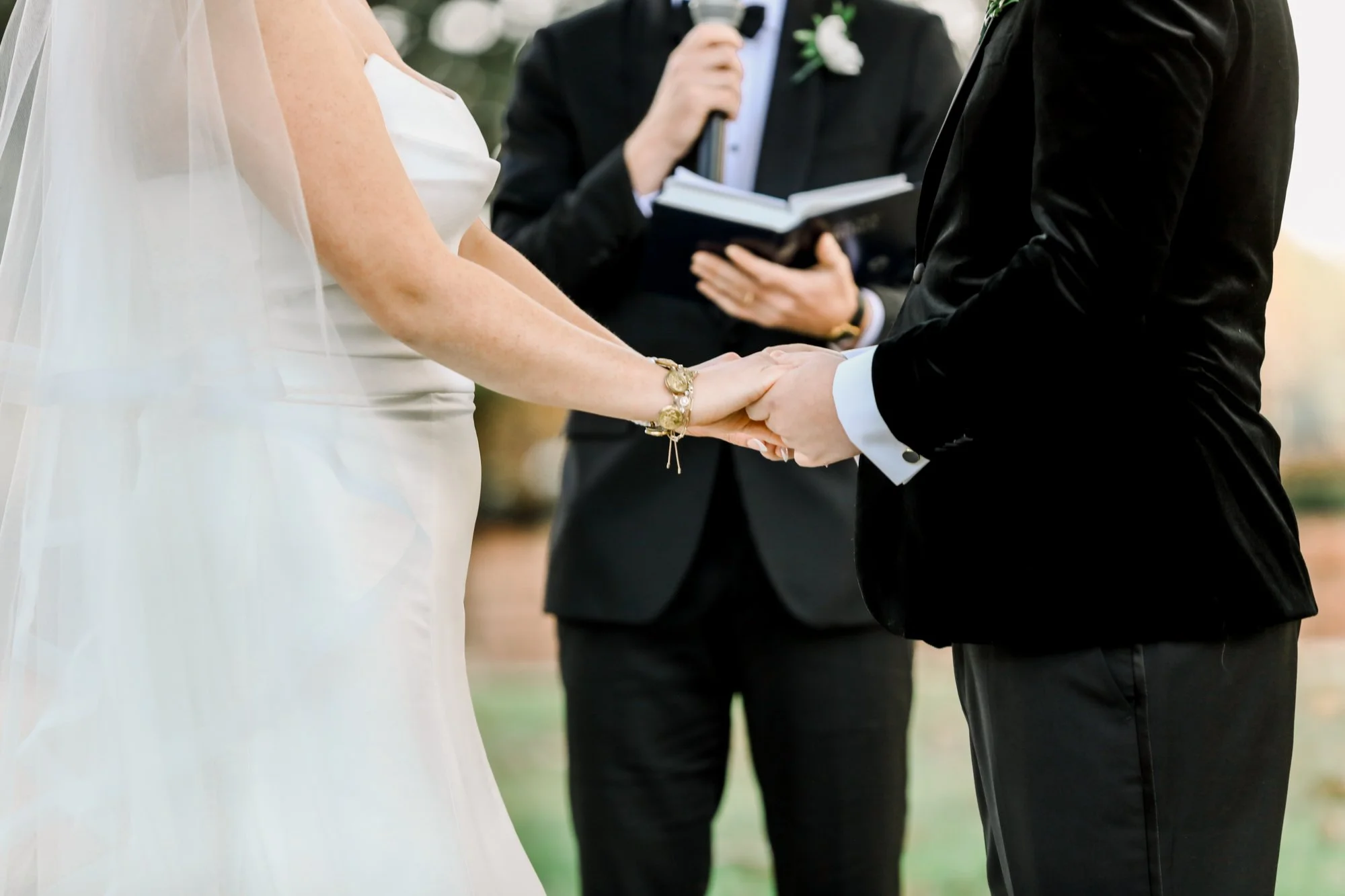 A bride and groom holding hands during their wedding ceremony, with an officiant standing behind them reading from a book.