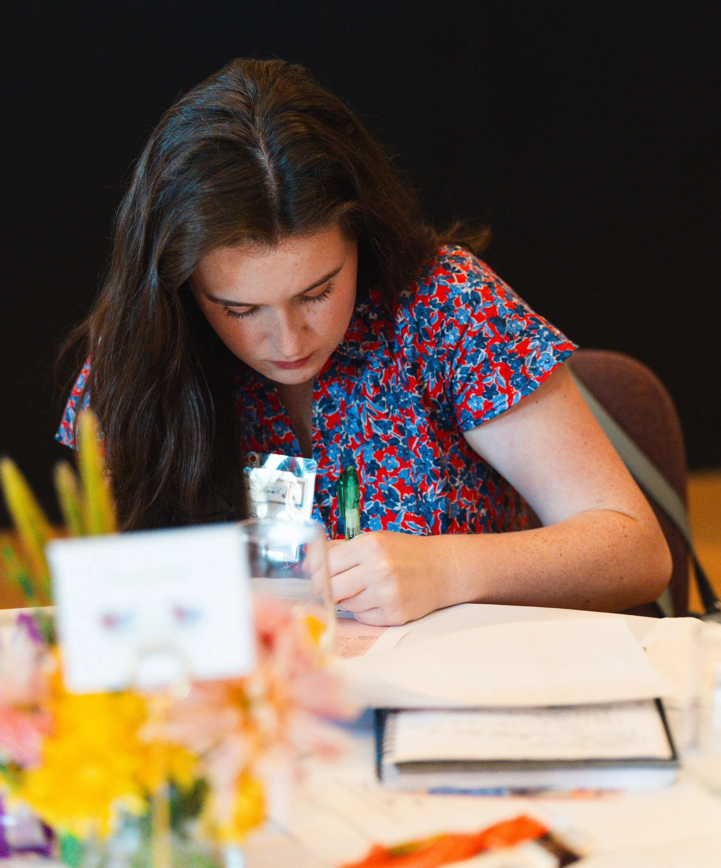 A young woman with long dark hair and freckles writing at a cluttered table with papers, notebooks, and blurred flowers in front of her.