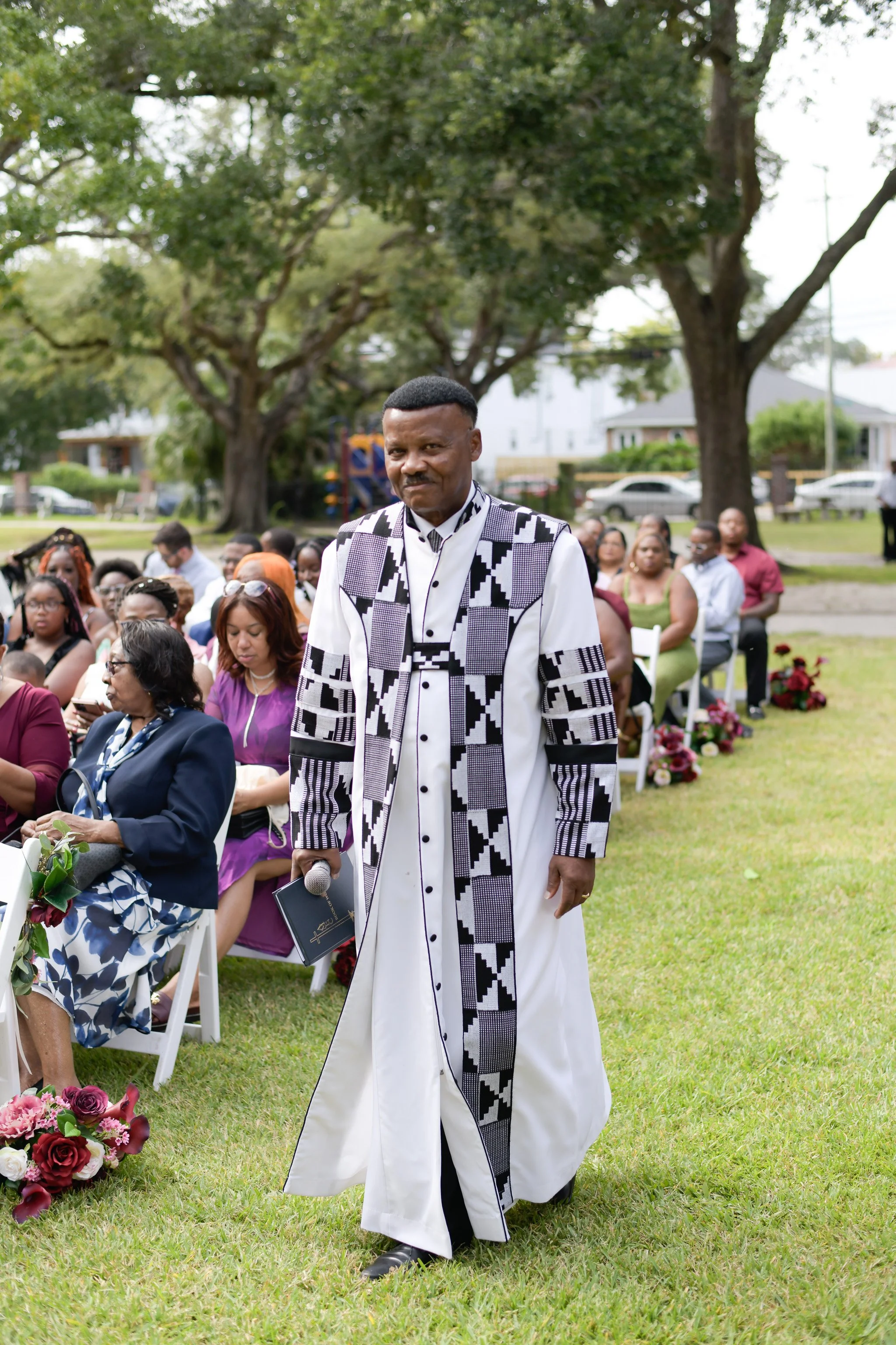 A man in a white and black patterned robe walking in front of seated guests at an outdoor event, with trees and houses in the background.