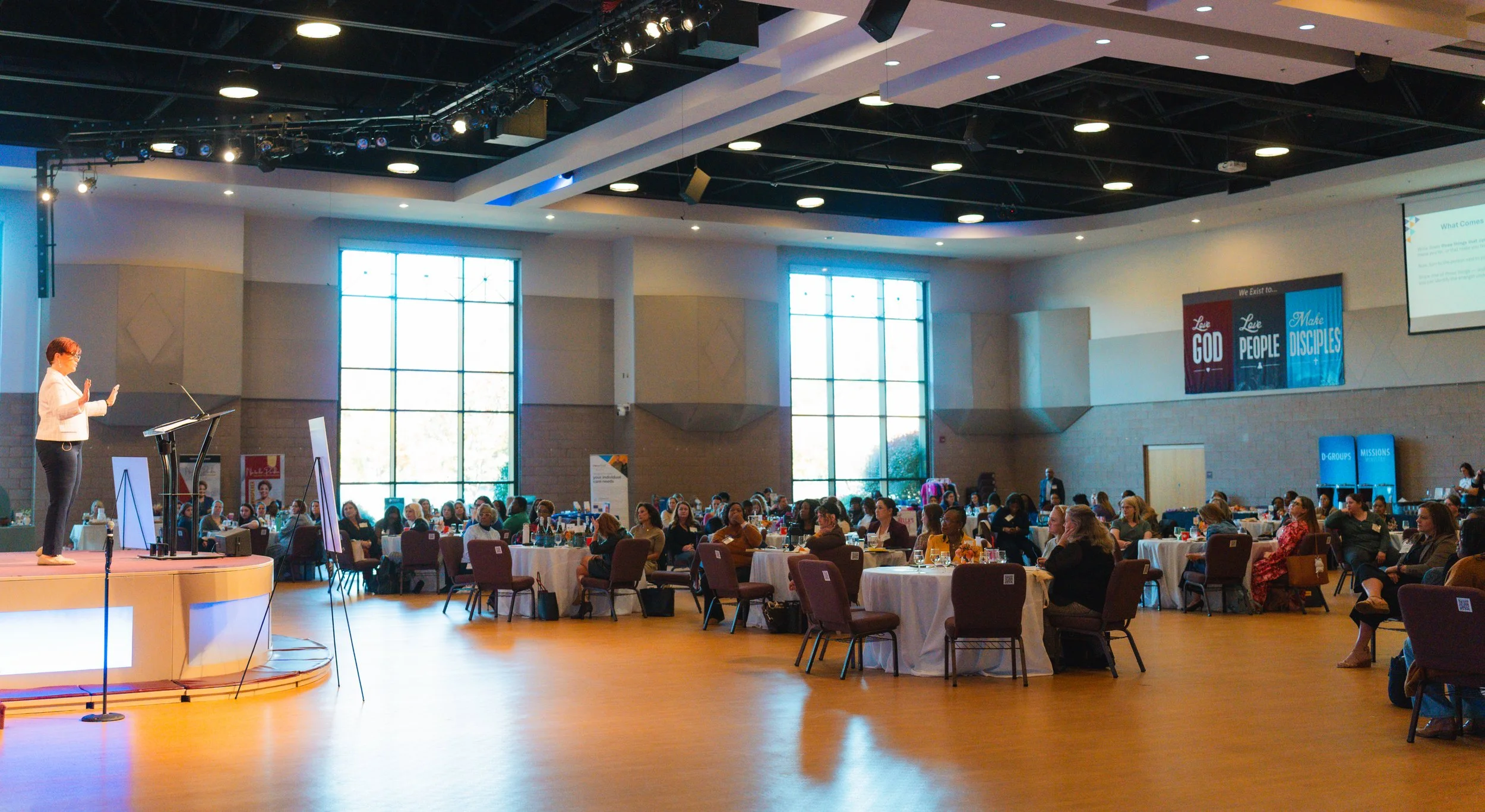A woman is speaking on stage at a conference, with an audience seated at round tables in a large, brightly lit auditorium.