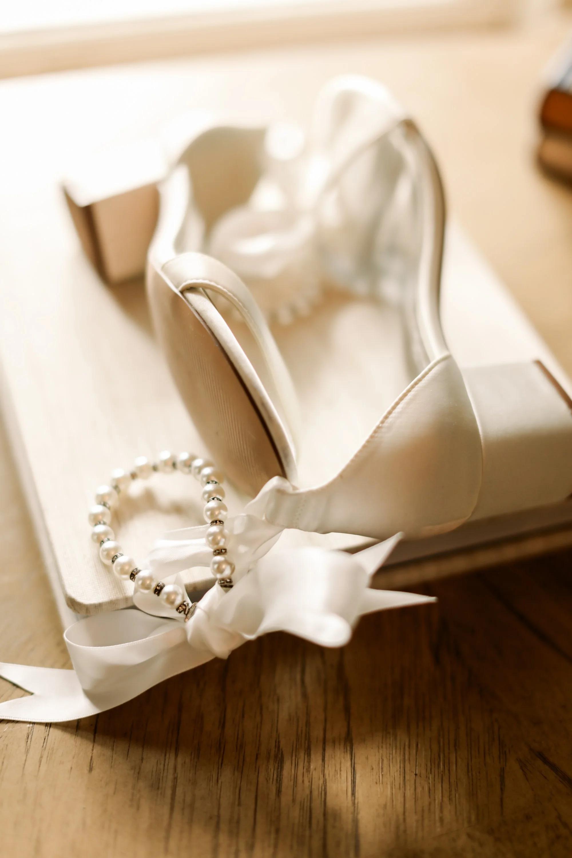 A white ribbon-tied pearl bracelet next to a small open box with a pair of white sneakers inside, placed on a wooden surface.