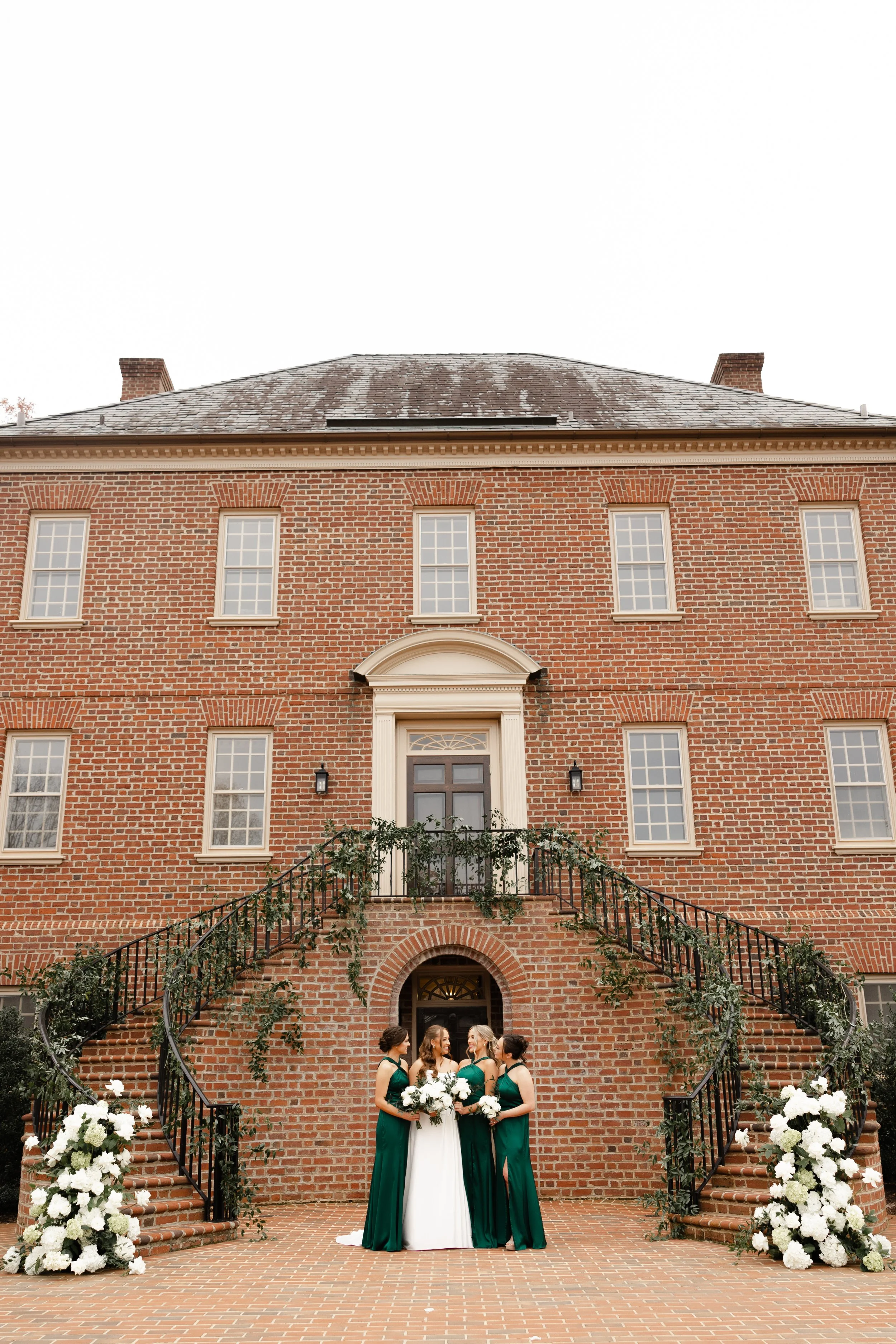 A bride in a white wedding dress holding a bouquet, surrounded by three bridesmaids in green dresses, standing in front of a brick building with an arched entrance and floral decorations.