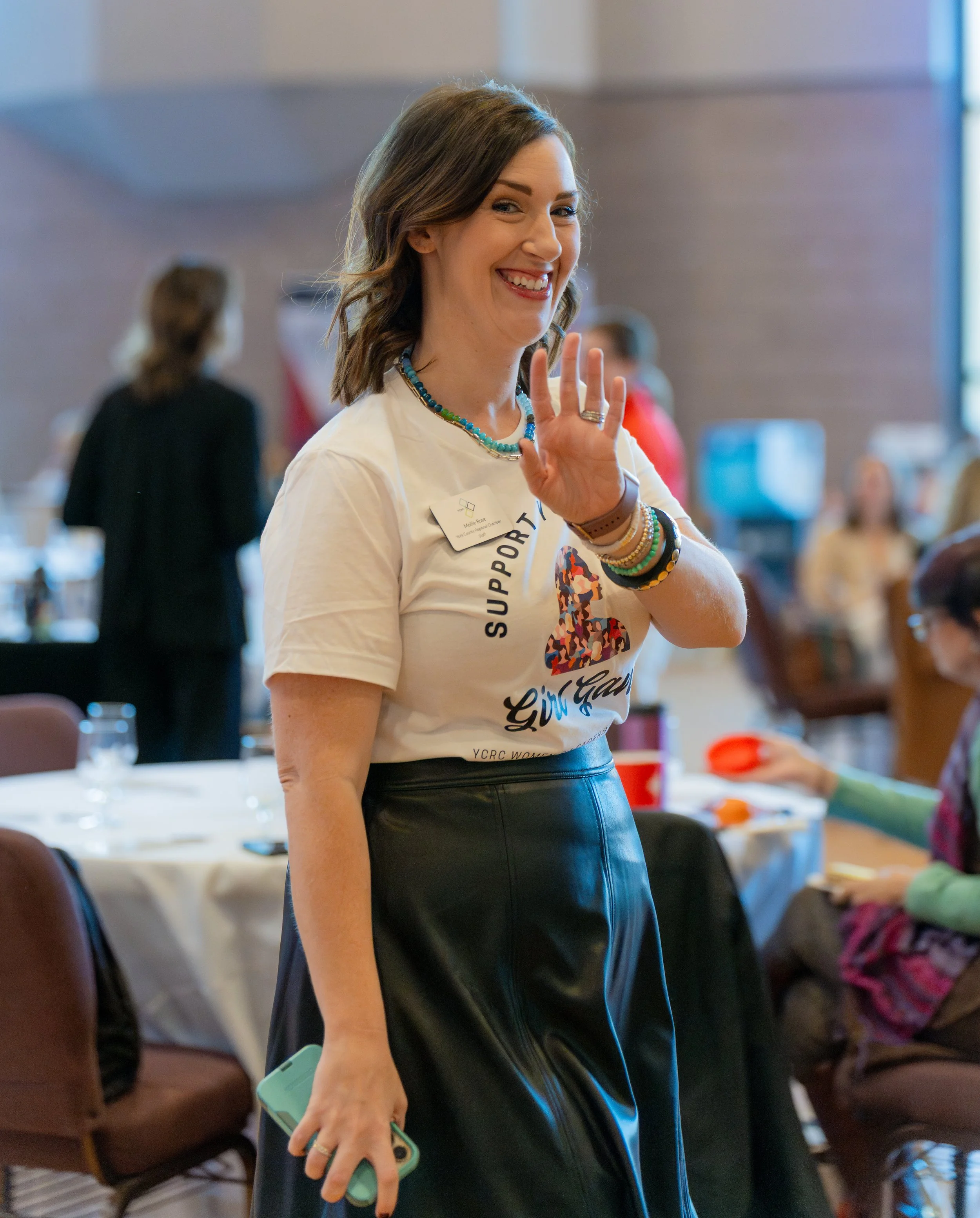 A woman smiling and waving at a conference or event, holding a phone with a mint green case, wearing a white shirt with event and supporter text, and multiple bracelets.