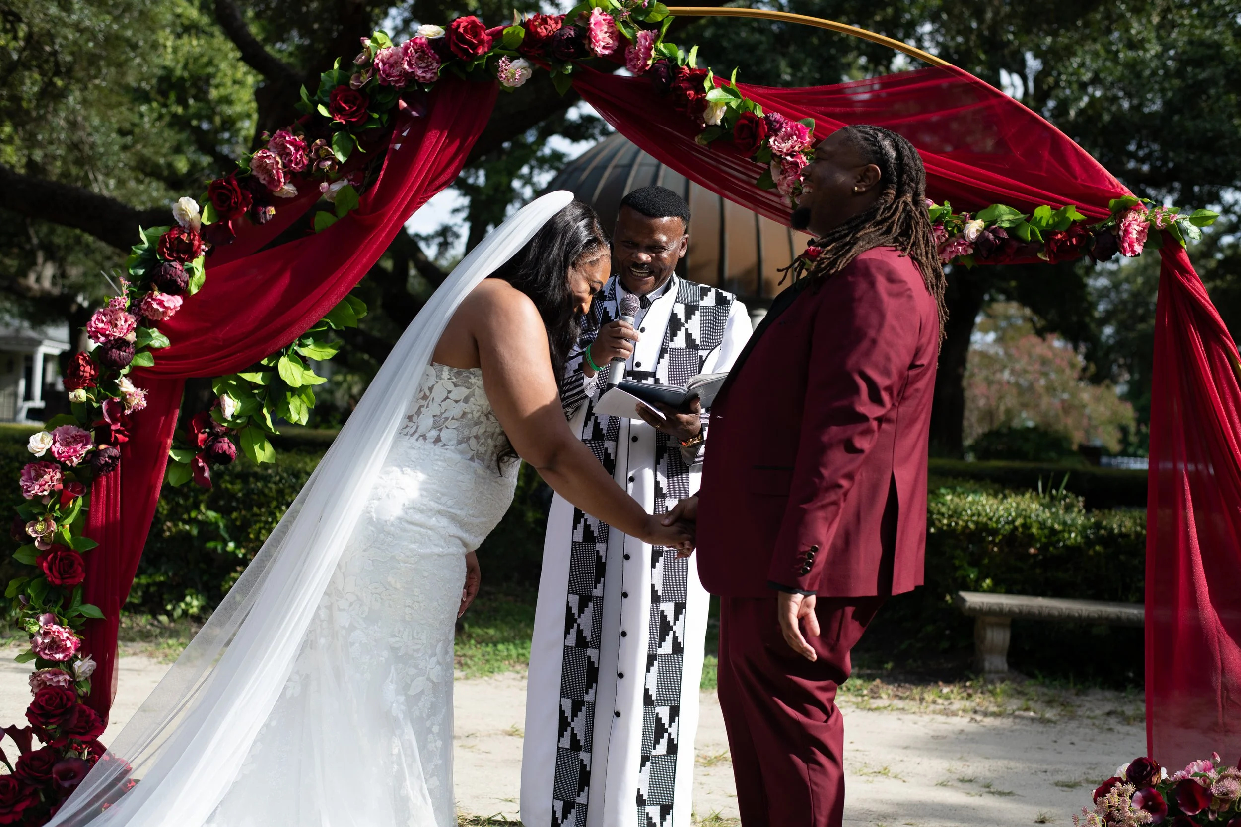 A couple getting married outdoors under a floral arch, with a minister officiating, on a sunny day with trees in the background.