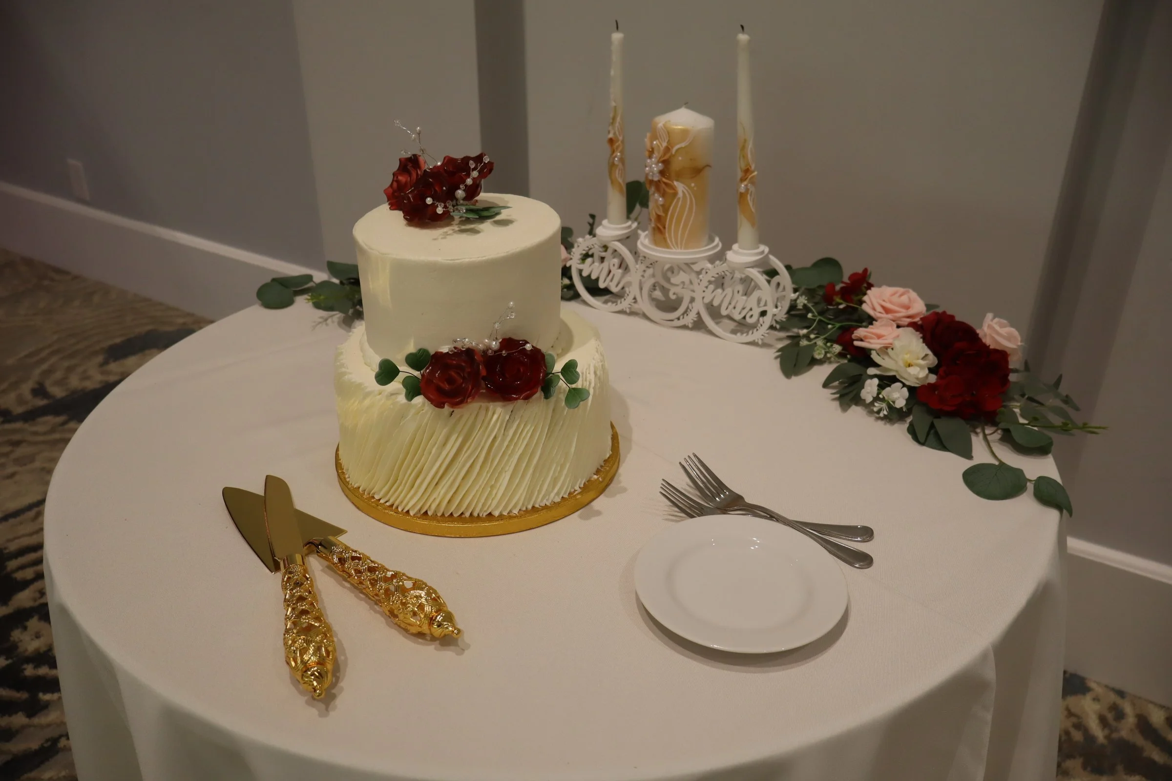 Two-tiered wedding cake decorated with red roses and green leaves, placed on a round table with a floral centerpiece, gold cake knives, a small white plate, and two forks, with a candleholder featuring three candles in the background.