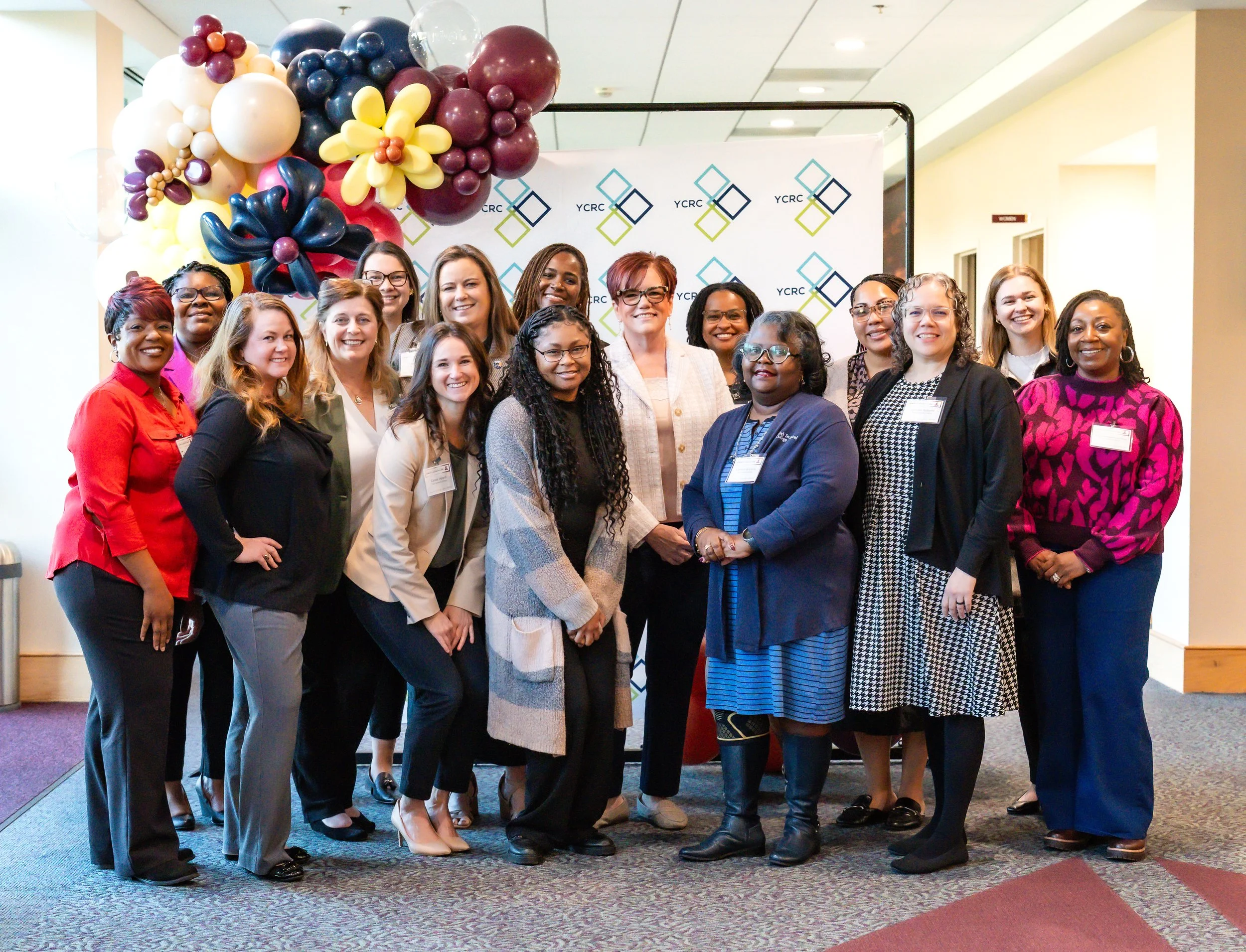 A diverse group of women posing for a photo at an event with a backdrop that reads 'YCRC' and decorated with oreo-shaped balloons in purple, gold, white, and black.