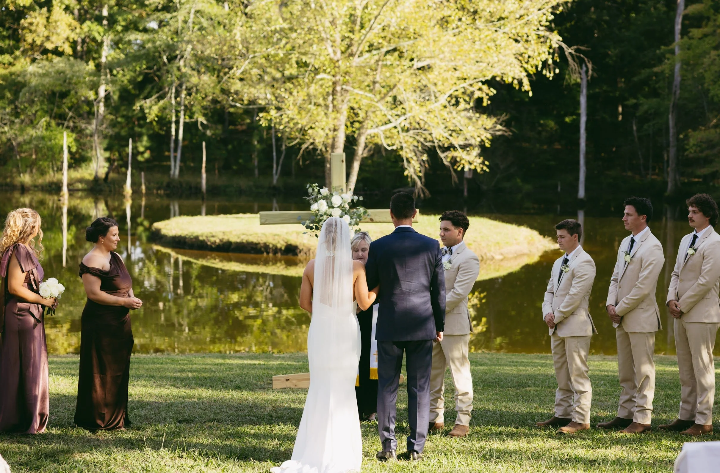 A wedding ceremony outdoors by a pond, with a bride and groom standing before a officiant, surrounded by bridesmaids and groomsmen in formal attire, with trees and water in the background.