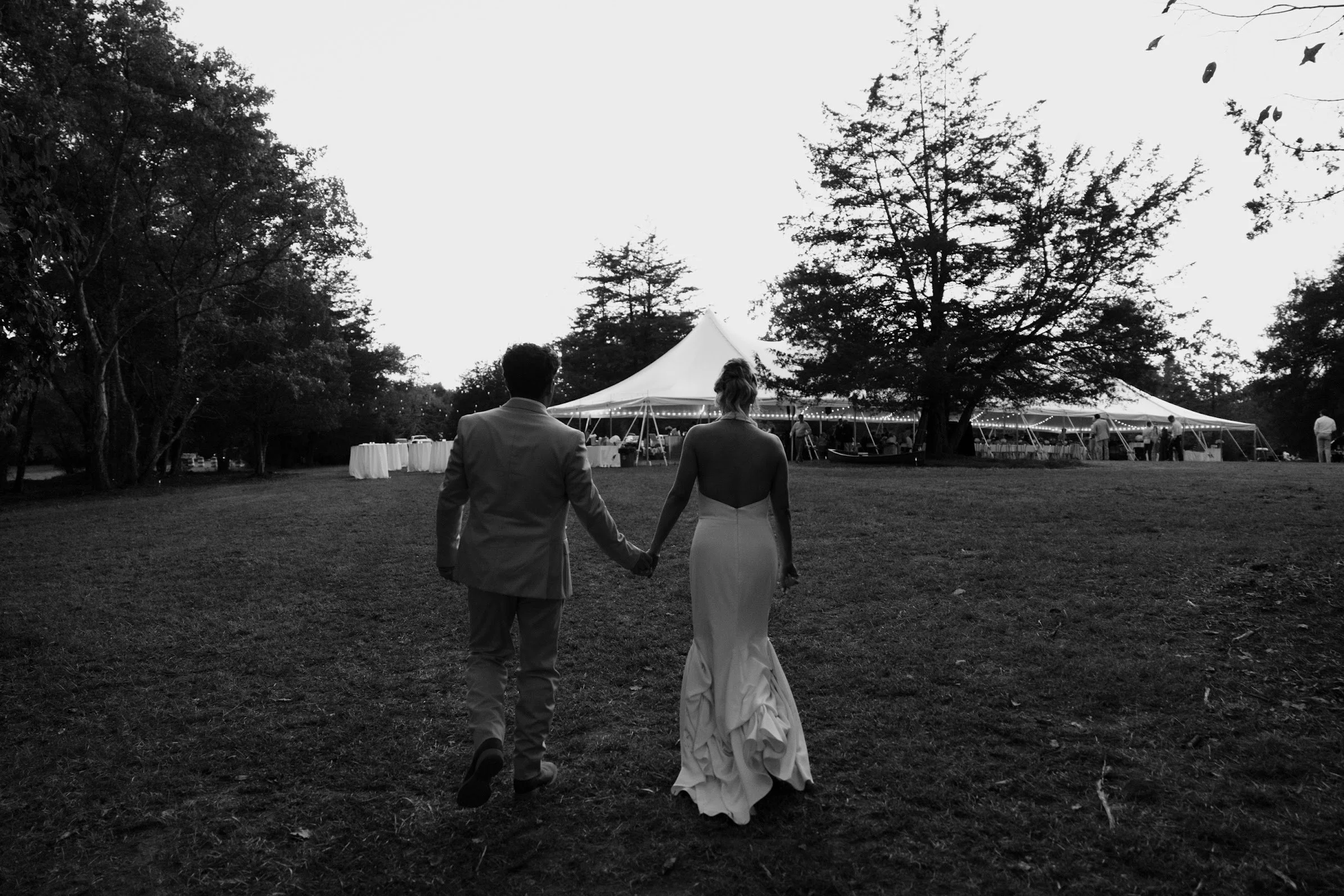 A couple dressed in wedding attire walking hand in hand towards a large outdoor tent at sunset, with trees surrounding the area.