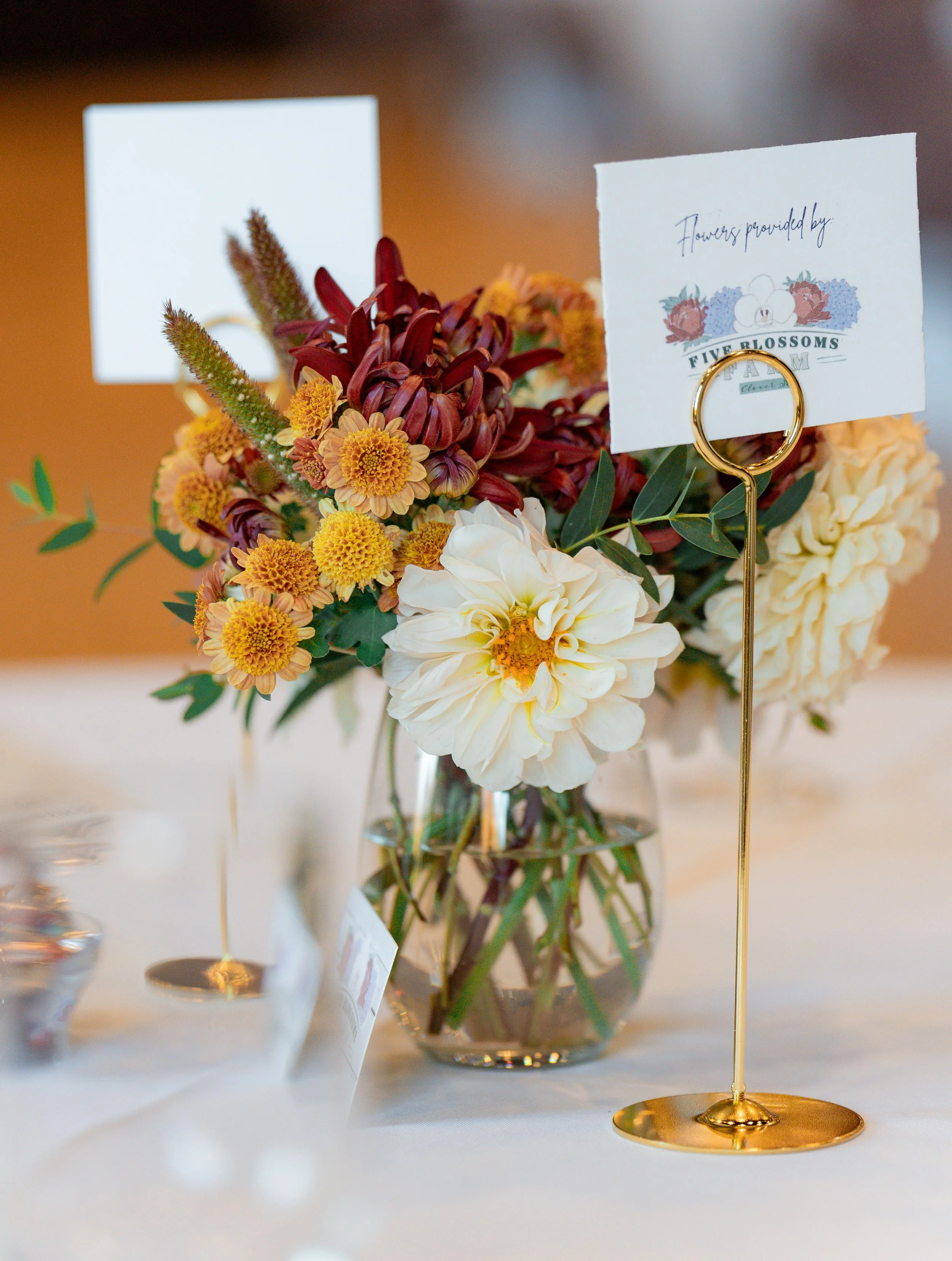 A floral centerpiece in a glass vase with various flowers, including white dahlias, yellow daisies, and red lilies, on a white table with a card and a gold ring holder.