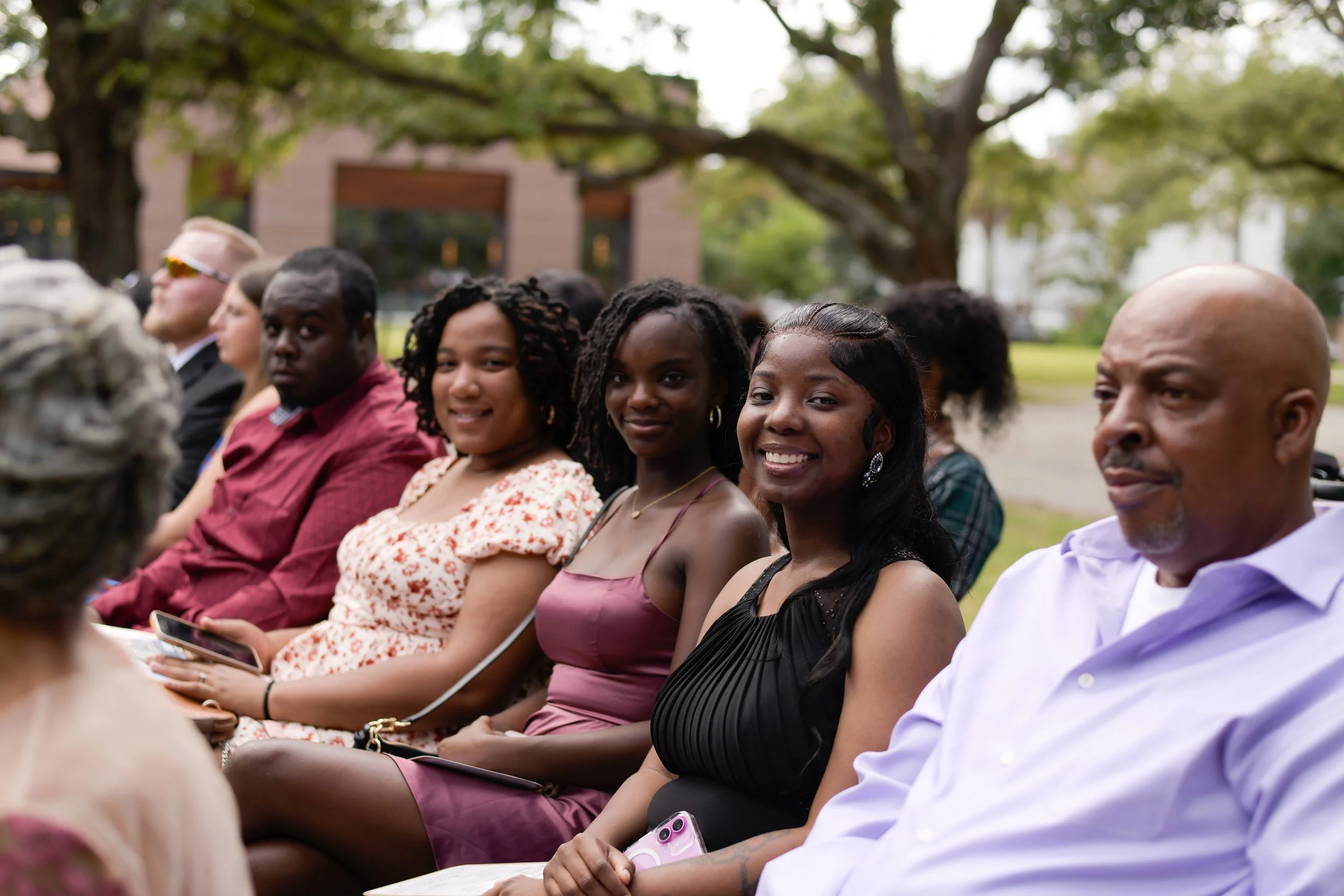 Group of people sitting outdoors during daytime, smiling and engaging at an event with trees in the background.