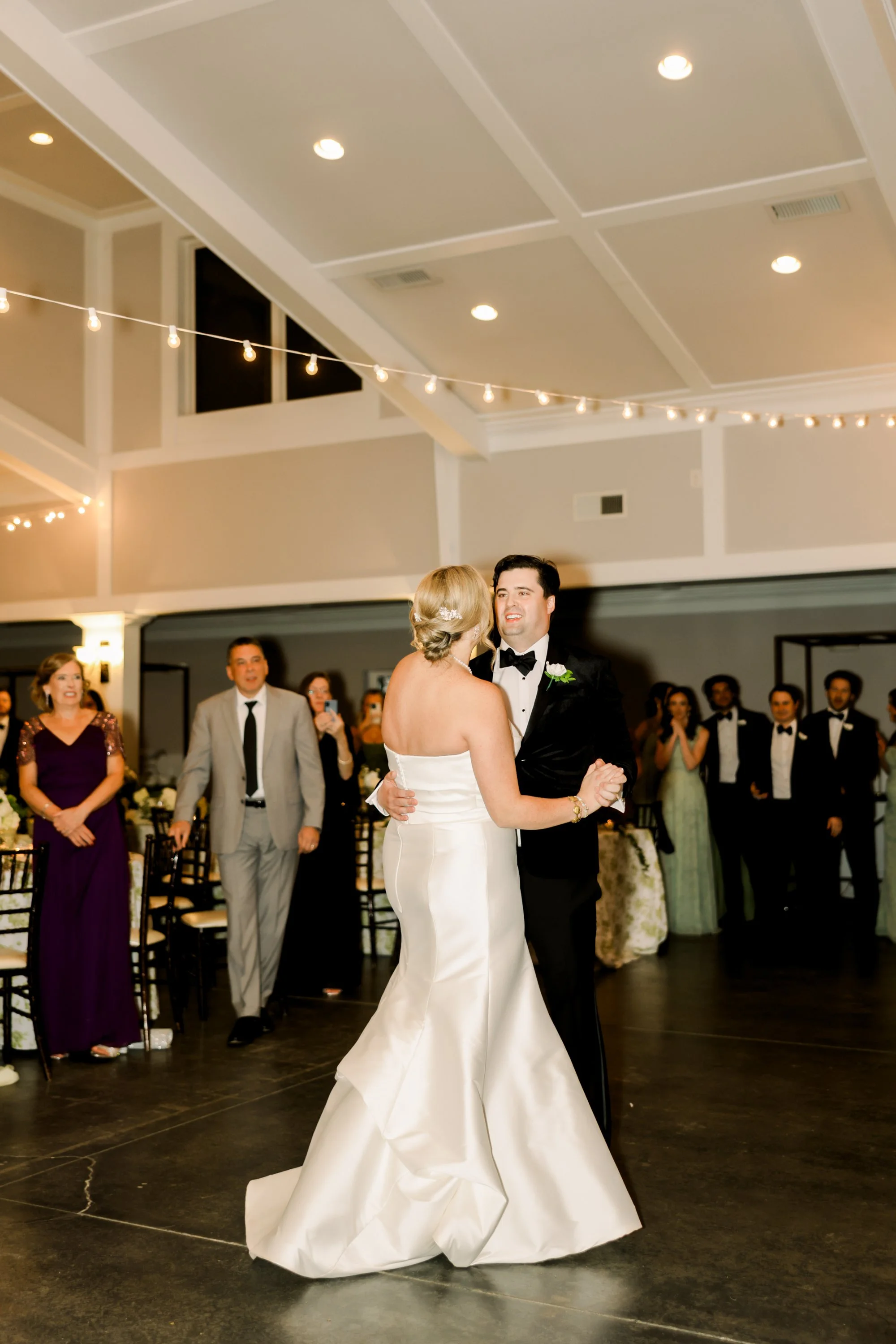 A bride and groom share their first dance at a wedding reception, surrounded by guests in formal attire in a decorated banquet hall.