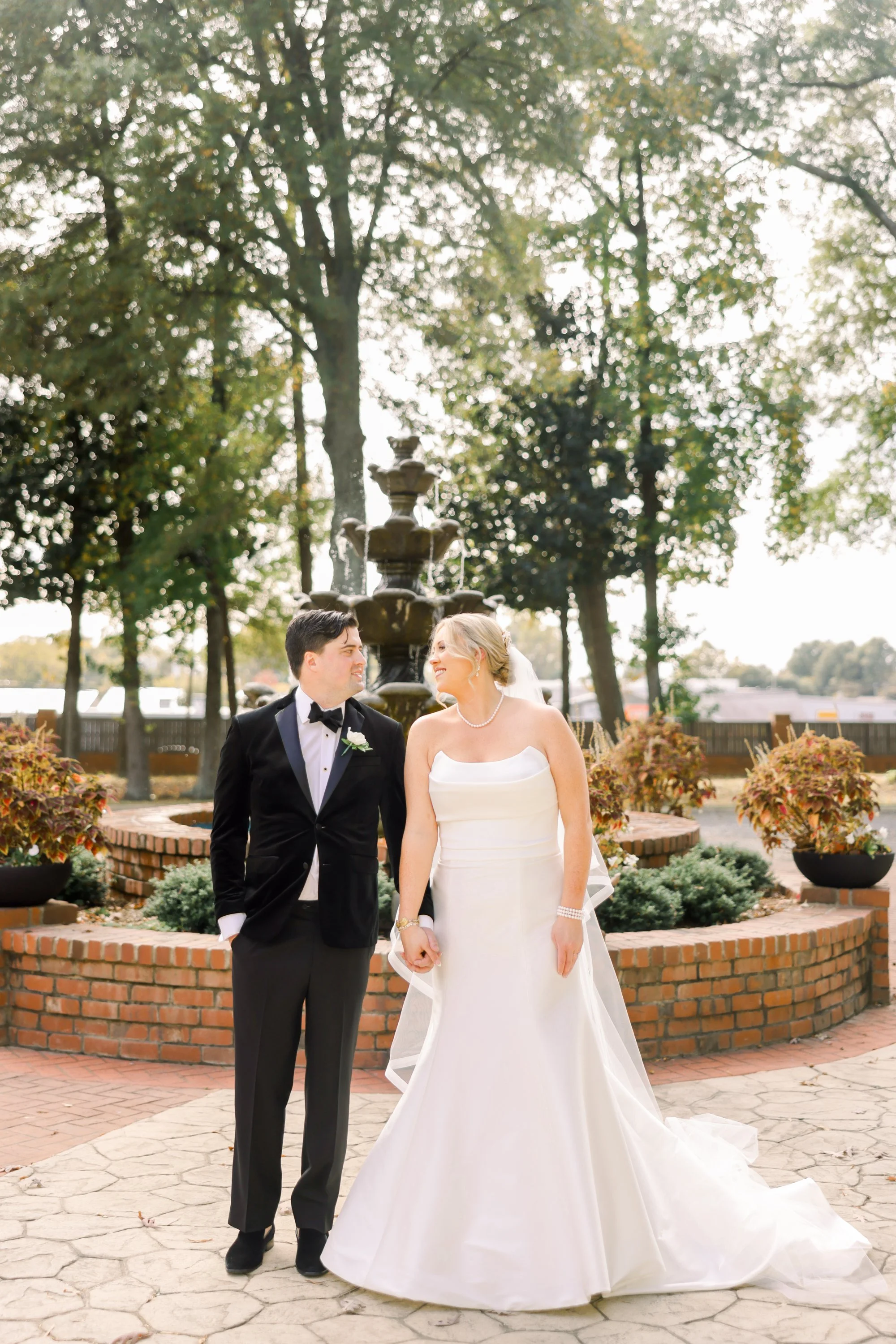 A bride and groom stand hand in hand outdoors near a fountain, smiling at each other on their wedding day. The bride wears a white strapless wedding gown and the groom a black tuxedo with a bowtie. Bright daylight and trees provide a scenic backgroun