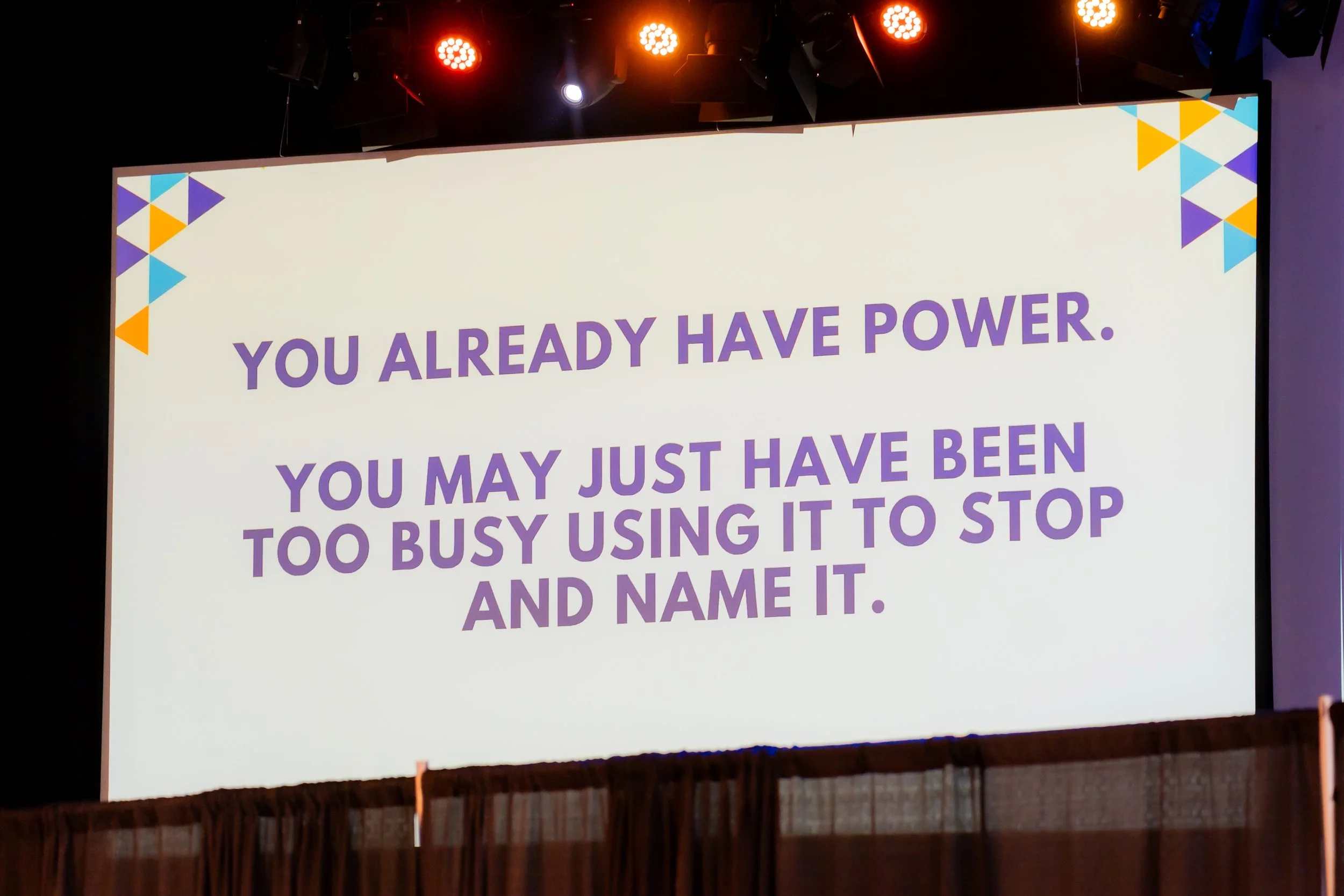 A large white screen displays an inspirational quote in purple text, reading: 'You already have power. You may just have been too busy using it to stop and name it.' The background includes colorful geometric shapes in the corners, with stage lightin