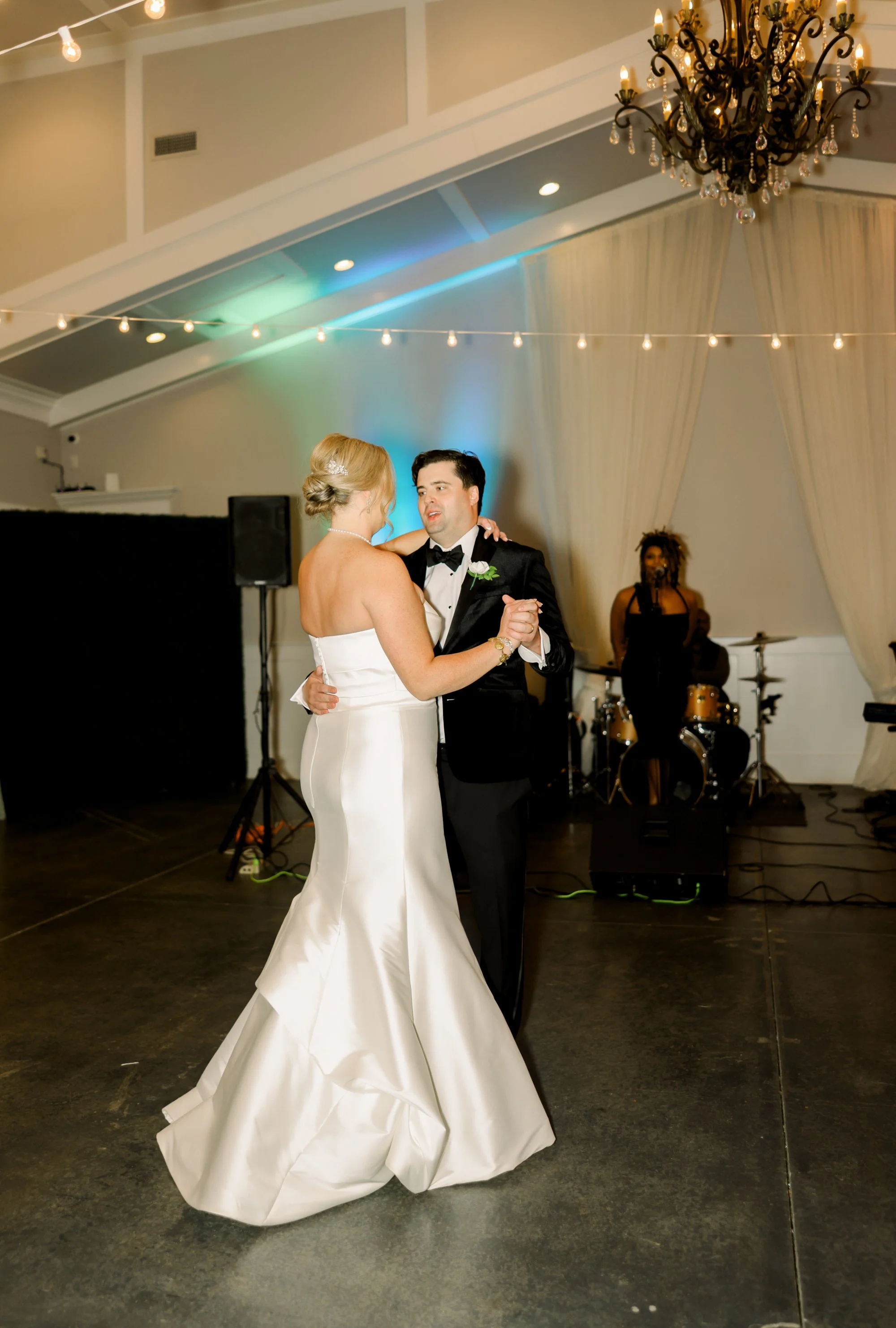 A bride and groom dance together at their wedding reception, with a live band playing in the background and string lights overhead.
