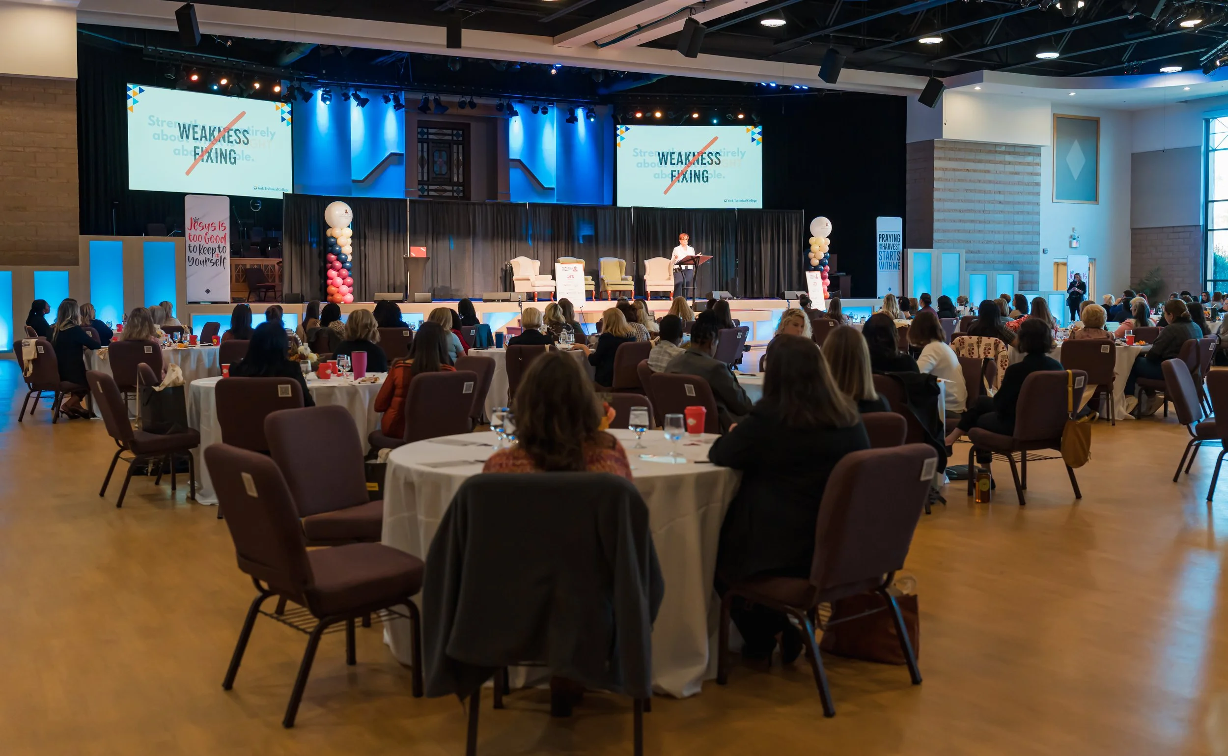A large conference room filled with round tables and chairs occupied by attendees listening to a speaker on a stage. The stage has screens displaying the word 'WEAKNESS' with a red slash through it, and there are balloons and banners with motivationa
