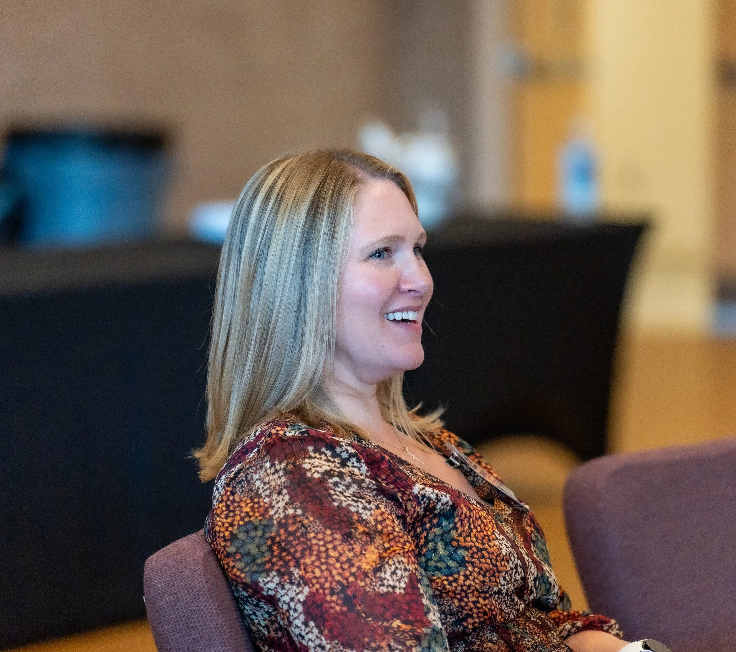 A woman with shoulder-length blonde hair, wearing a colorful patterned blouse, smiling and sitting on a chair in an indoor setting.