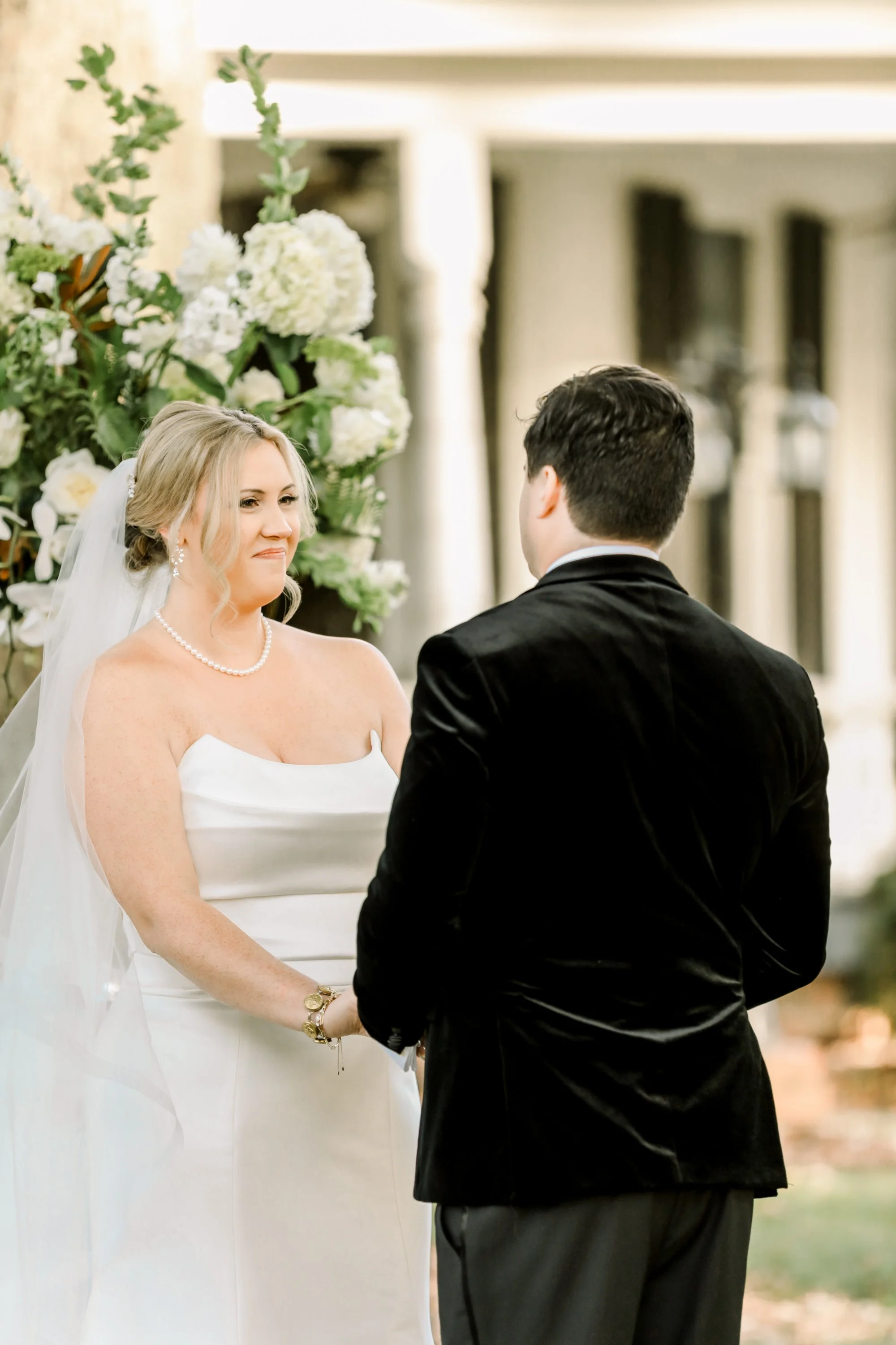 A bride and groom exchange vows outdoors with a floral backdrop.