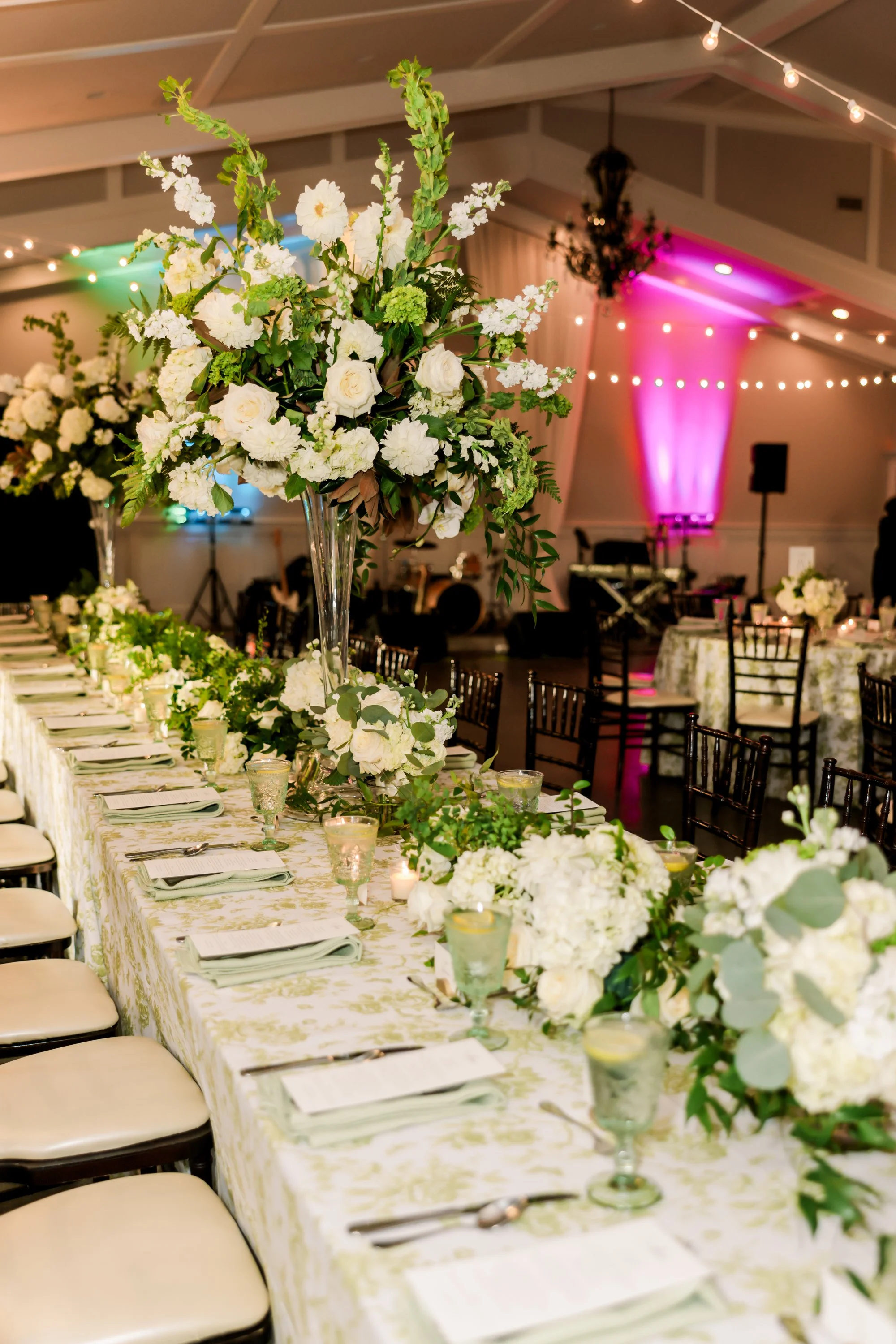 Elegant banquet table decorated with white flowers in tall vases, candles, and place settings, set up for a celebration or wedding in a tent with string lights and colorful uplighting.