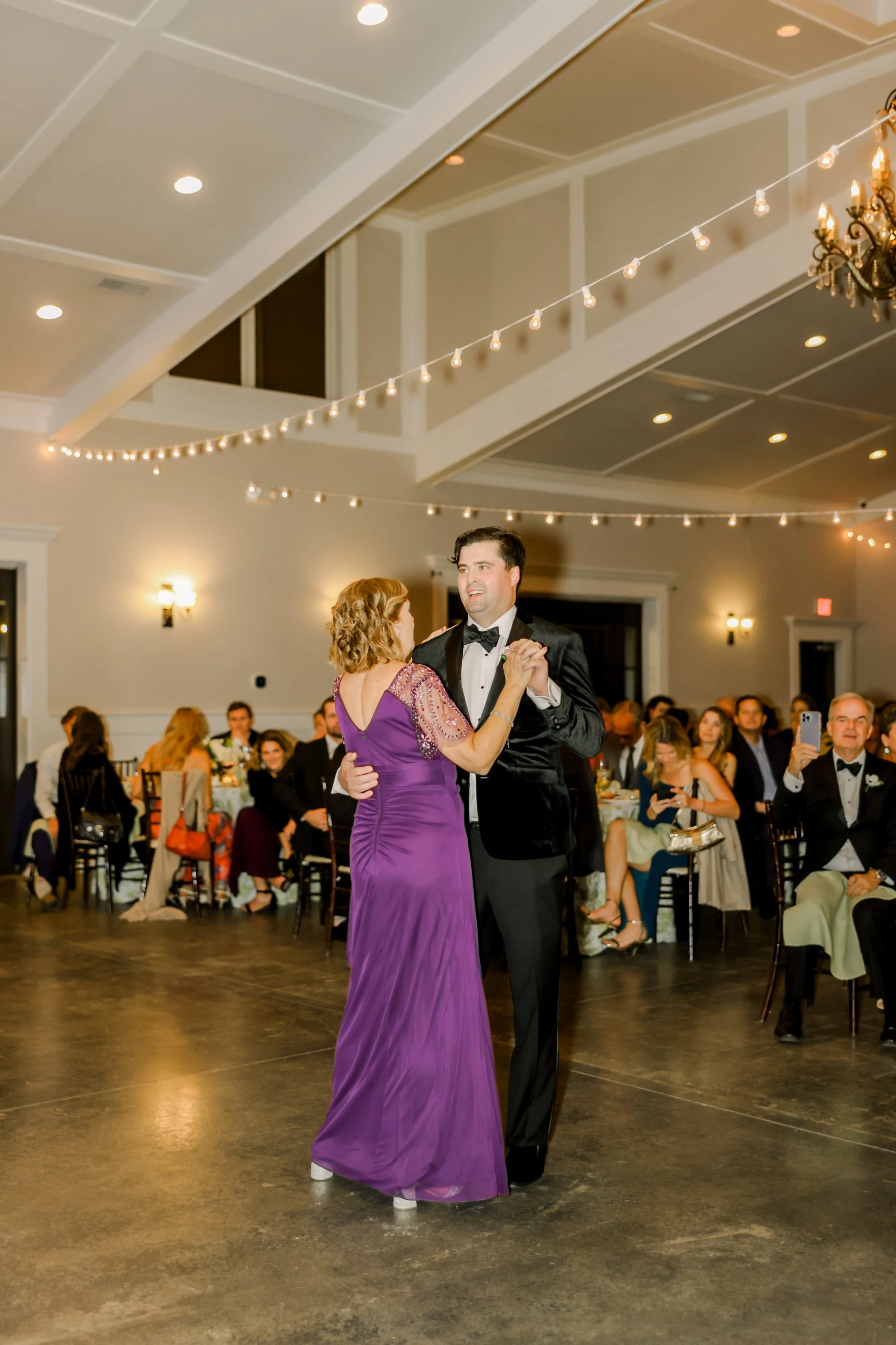 A couple is dancing at a formal event, with the man wearing a black tuxedo and the woman wearing a purple gown. They are surrounded by seated guests in an elegantly decorated hall with string lights and chandeliers.