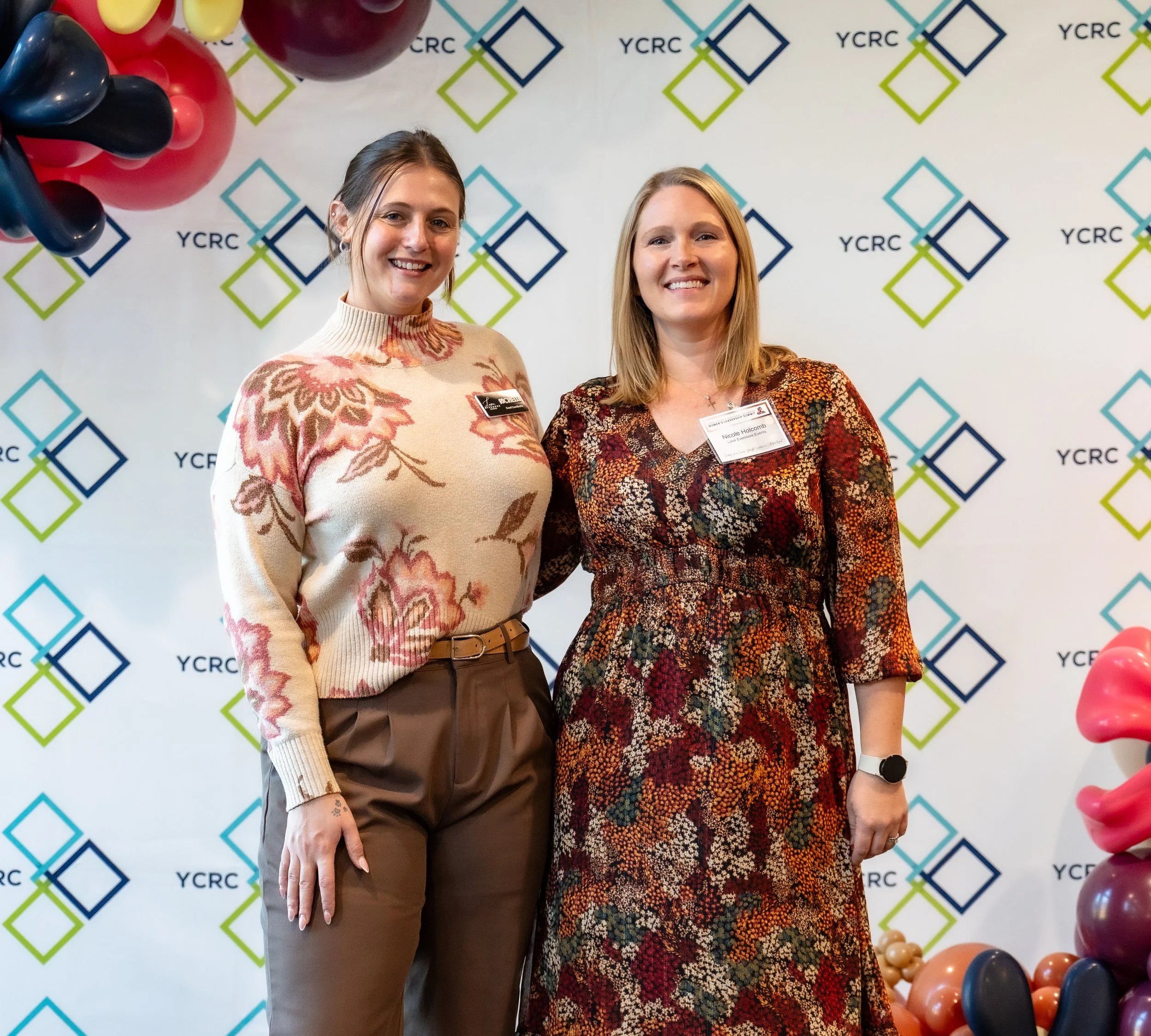 Two women standing smiling in front of a backdrop with YCRC logos, one wearing a floral sweater and brown pants, the other wearing a floral dress with a name tag.