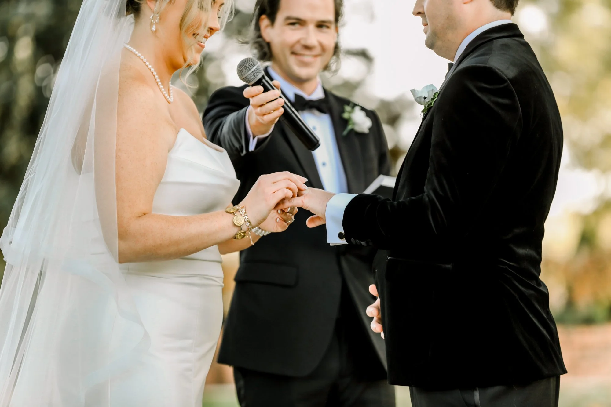 A bride and groom exchanging rings during their outdoor wedding ceremony, officiant holding a microphone, with trees and a blurred background.