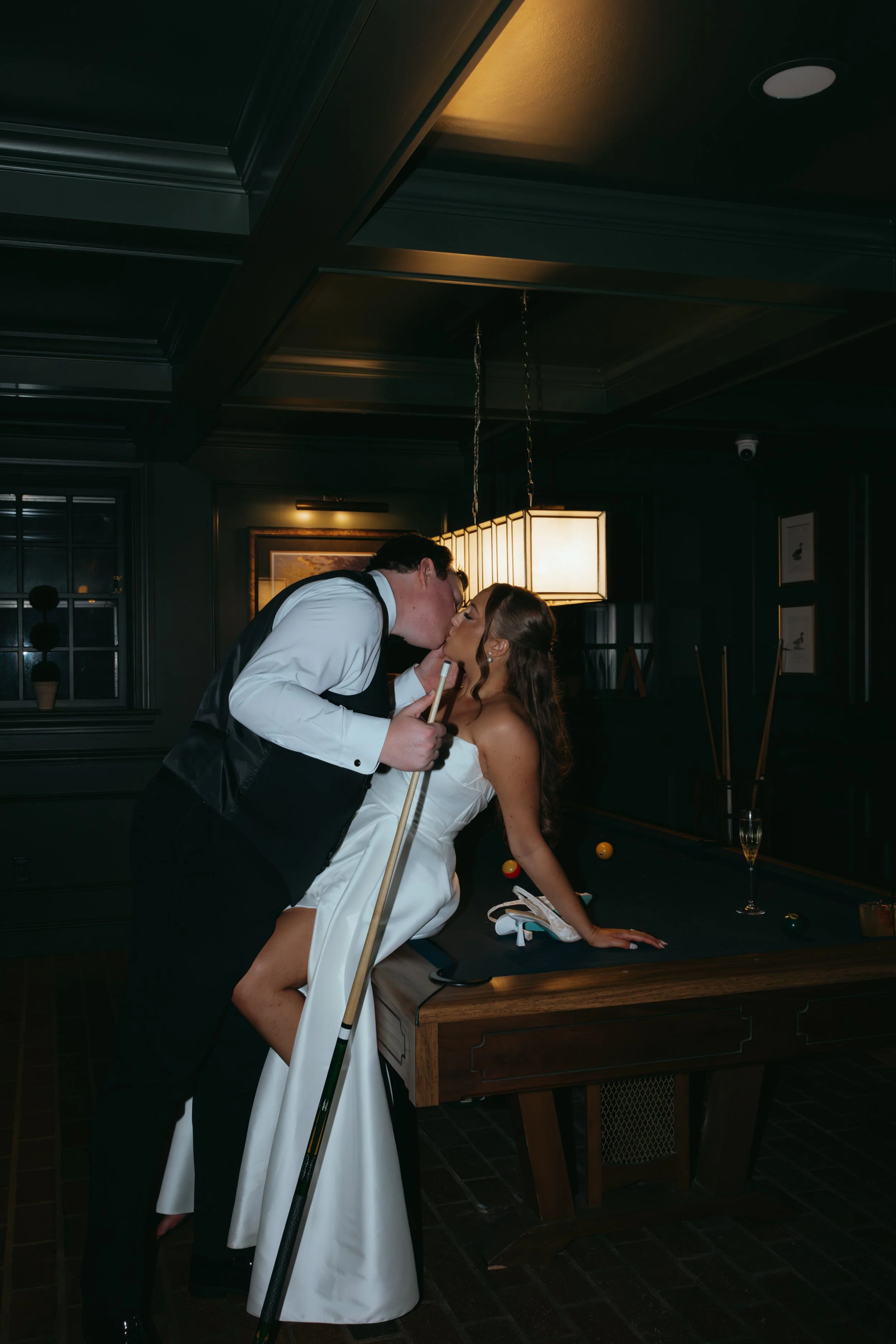 A man in formal attire kissing a woman in a wedding dress at a pool table in a dimly lit room.