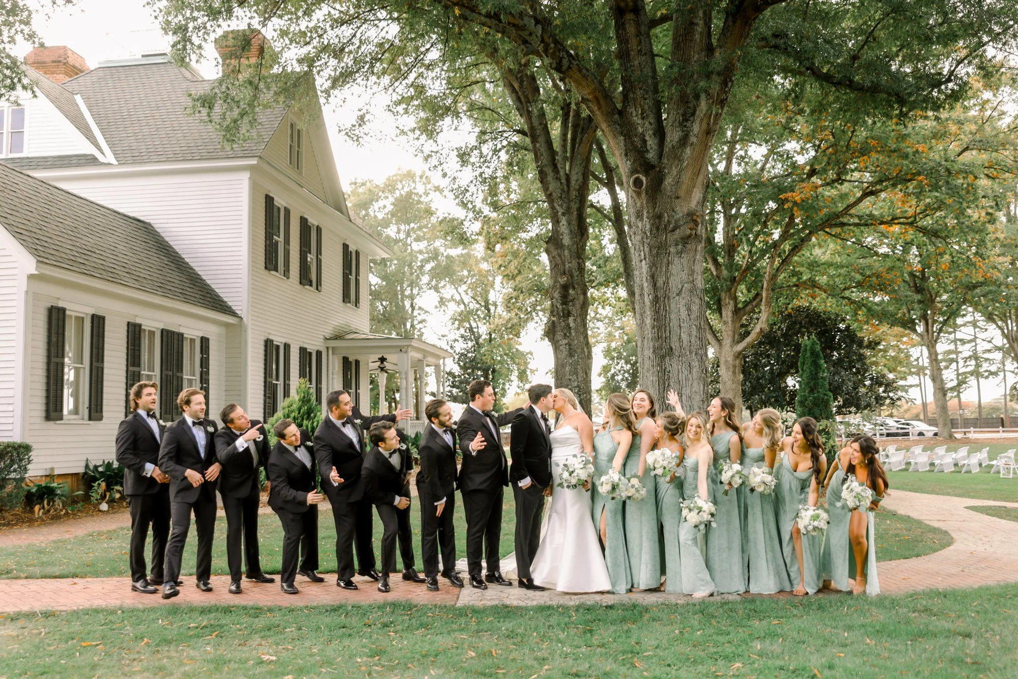 A wedding party of fourteen people standing outside in front of a large tree and a white house. The bride and groom are kissing in the center, with bridesmaids in light green dresses holding bouquets on the right and groomsmen in black suits on the l