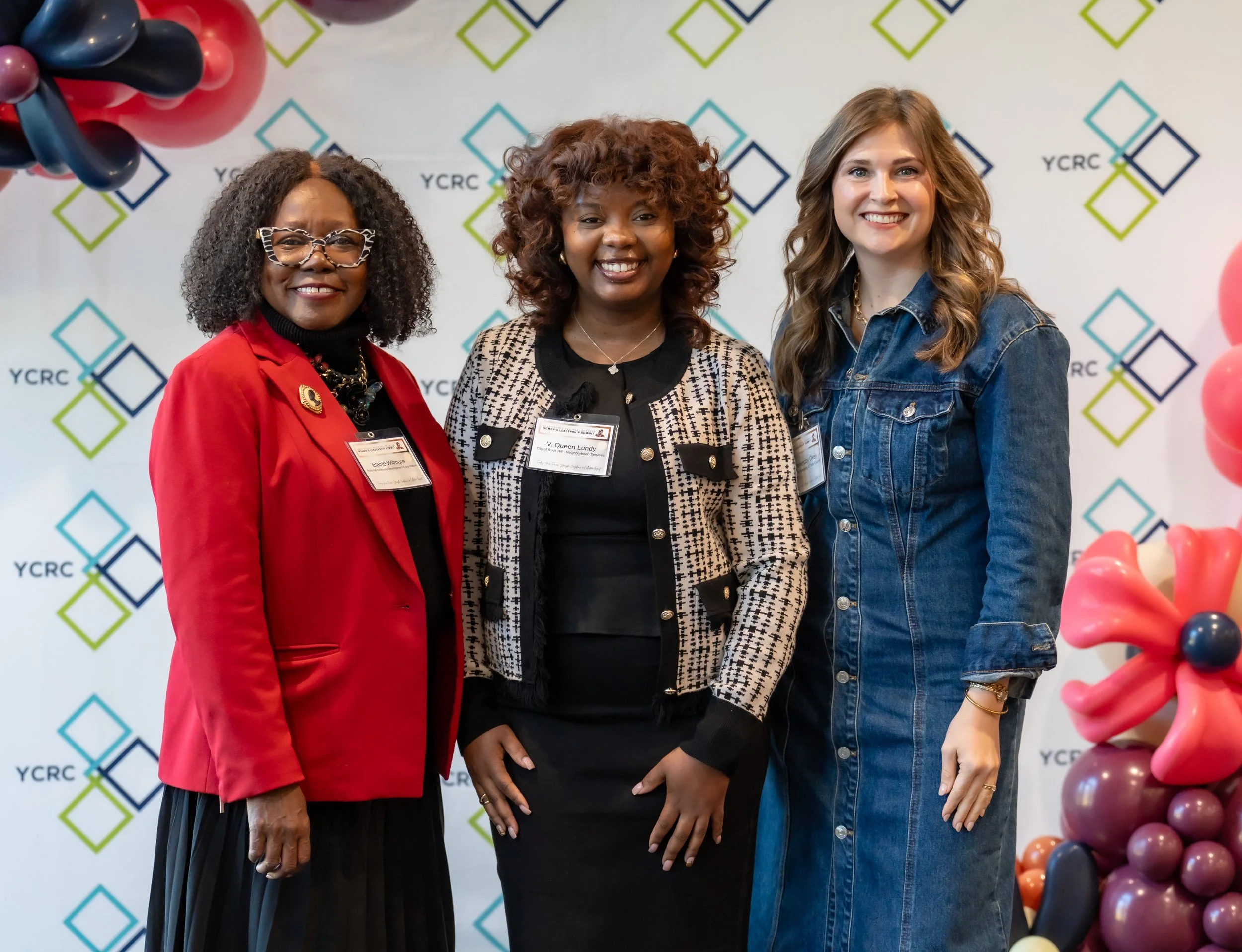 Three women standing together at a professional event, smiling at the camera. They are wearing name tags and attire suitable for a conference or seminar, with a decorated background featuring the YCRC logo and colorful balloon arrangements.
