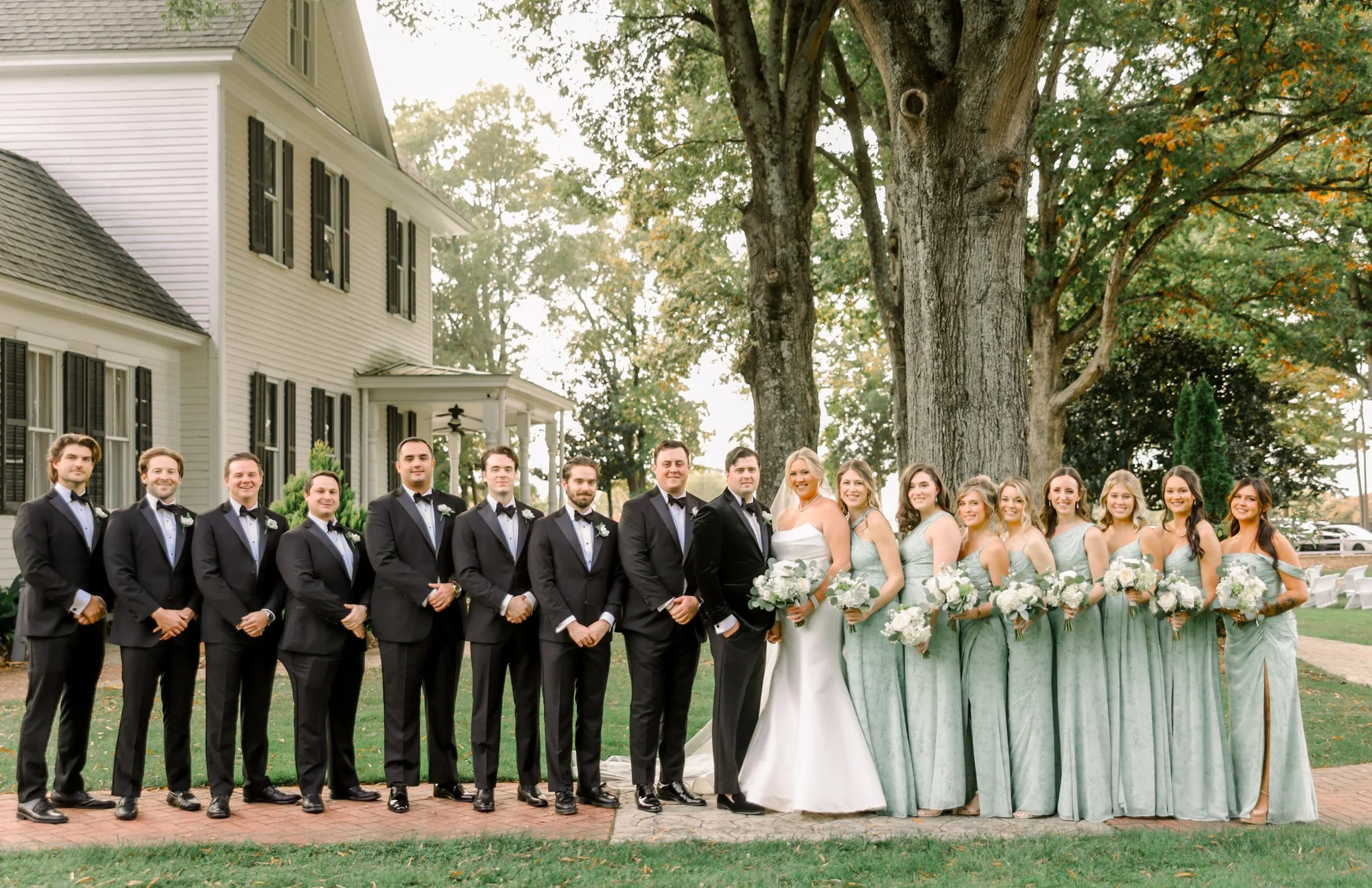 Group of wedding party members standing outdoors, dressed in formal attire, with bridesmaids in mint green dresses holding white bouquets and groomsmen in black tuxedos with bow ties, in front of a large tree and white house.