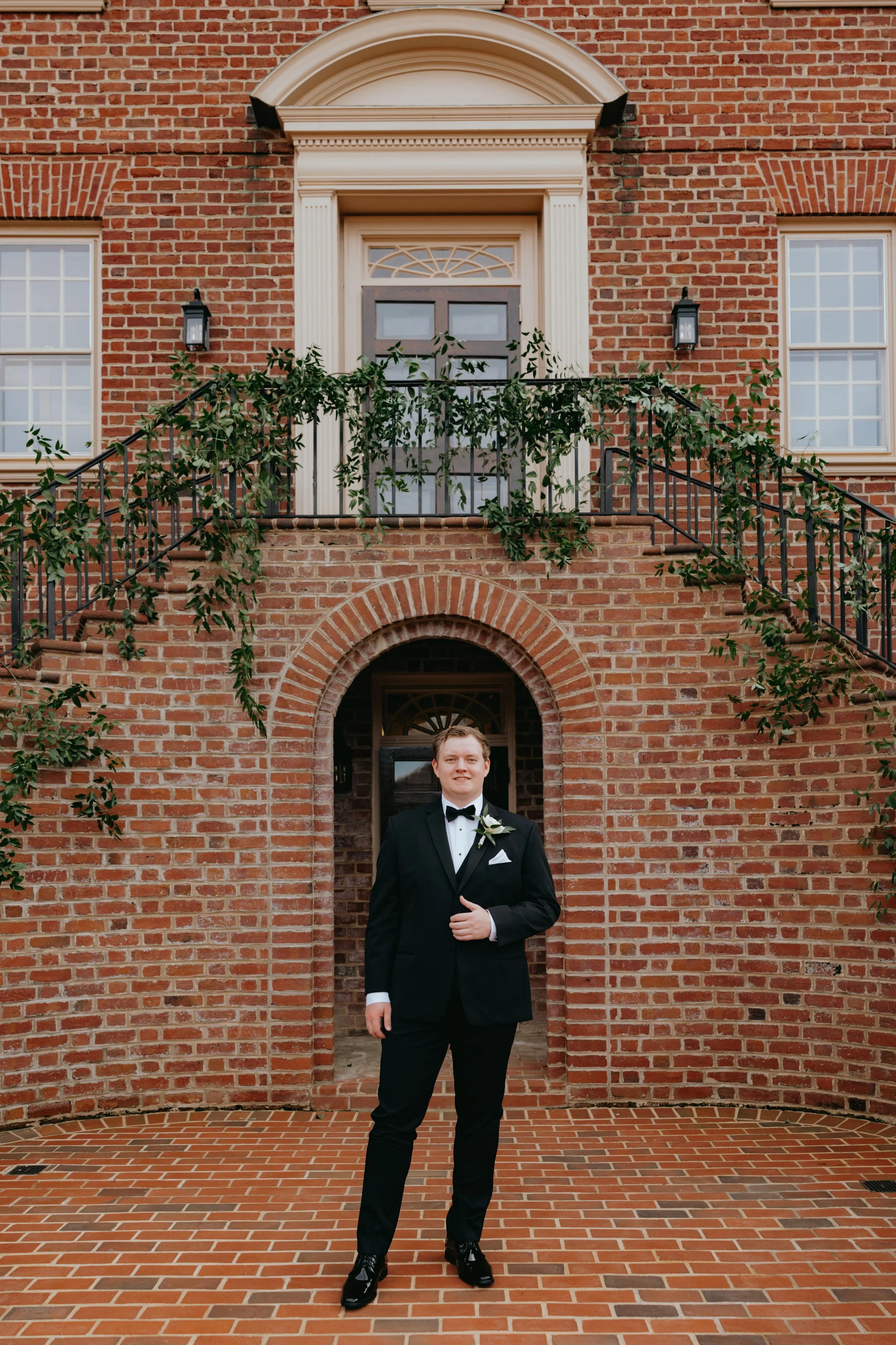 A man in a black tuxedo with a white shirt, black bow tie, and boutonniere, standing in front of a brick building with stairs and black railing, under an arched doorway.