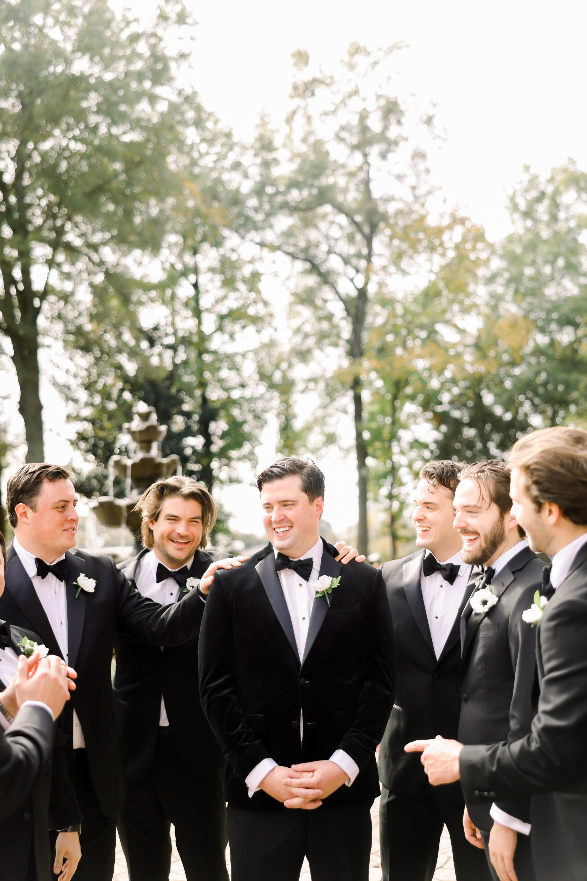 A group of men in tuxedos, gathered outdoors in a circle, smiling and congratulating a man in the center, at a wedding or formal event.