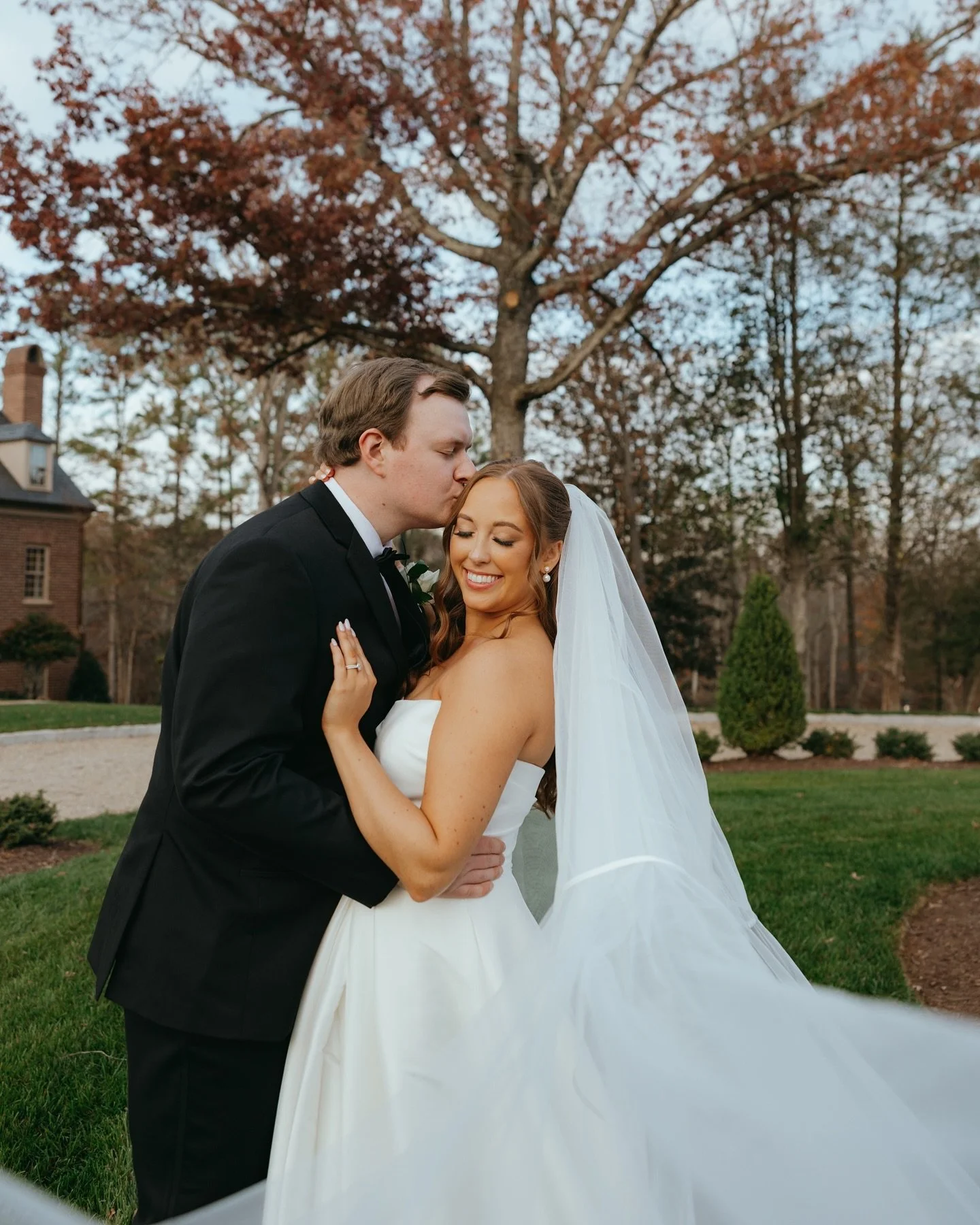 A bride and groom share an embrace outdoors during their wedding, with the groom kissing the bride on her forehead. The bride, wearing a white wedding gown and veil, smiles happily. The background features a large tree with autumn leaves and a house.