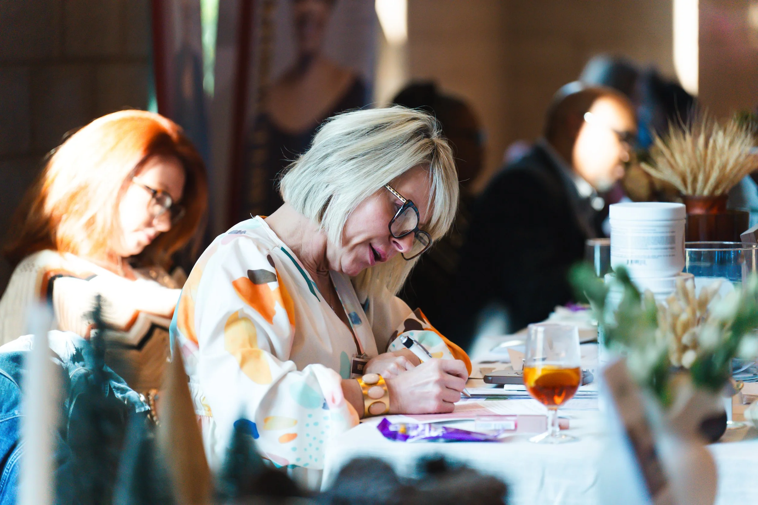 Woman with blonde hair and glasses writing on paper at a table, with a drink in front of her, in a well-lit room with other people in the background.