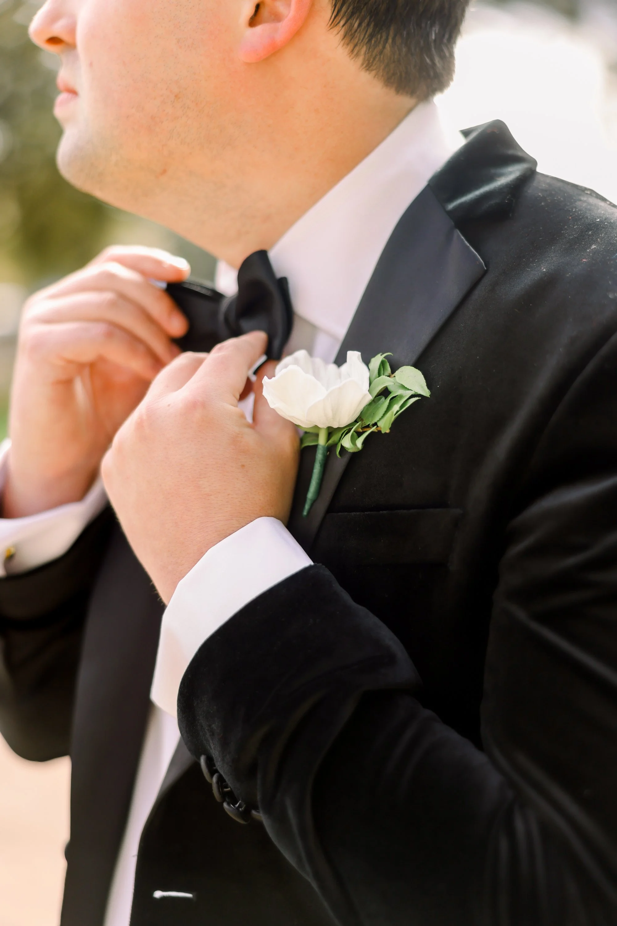 Man in tuxedo adjusting his bow tie, with a white flower boutonniere on his lapel.
