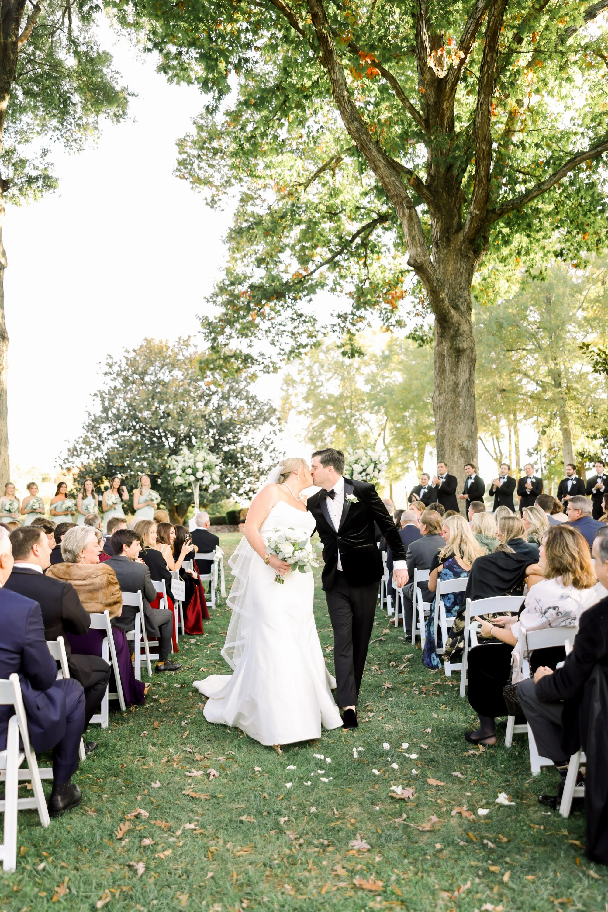 A bride and groom kiss under a large tree at an outdoor wedding ceremony. Guests seated on white chairs watch, with bridesmaids and groomsmen standing behind in the background.