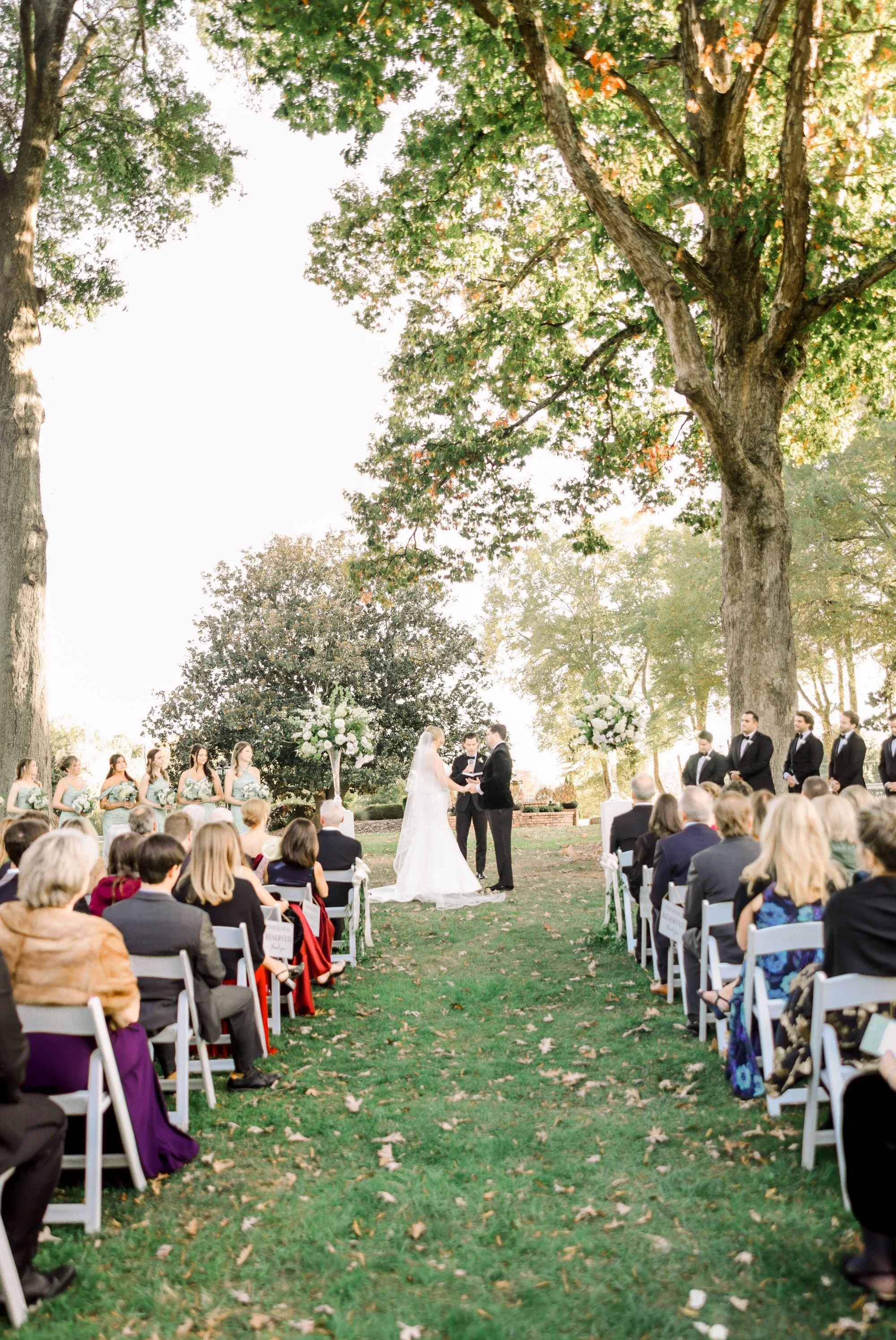 Outdoor wedding ceremony with bride and groom exchanging vows under large trees, surrounded by guests, bridesmaids, and groomsmen, in a scenic garden setting during daytime.